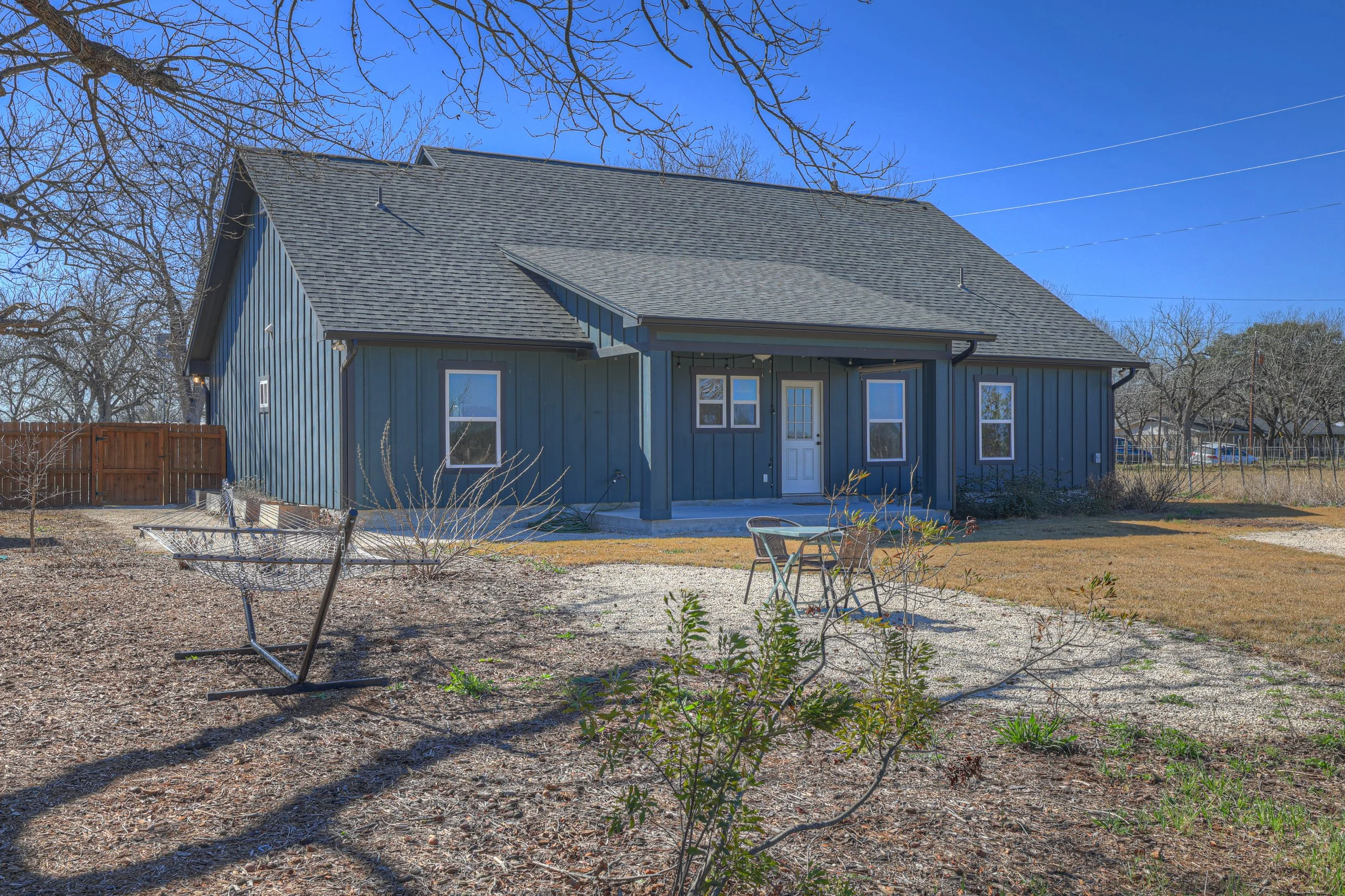 A blue house with a gray shingle roof, front porch, and white window frames. The yard has a gravel area with a metal table and chairs, a few small plants, and leafless trees in the background against a clear blue sky.