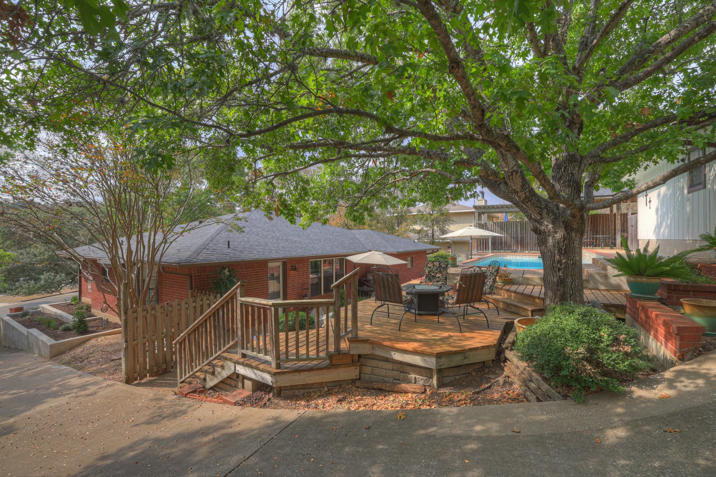 A backyard outdoor patio with a large tree providing shade over a wooden deck area. The deck has a table with four chairs, an umbrella, and a small staircase leading down to a concrete surface. In the background, there is a swimming pool with a paras