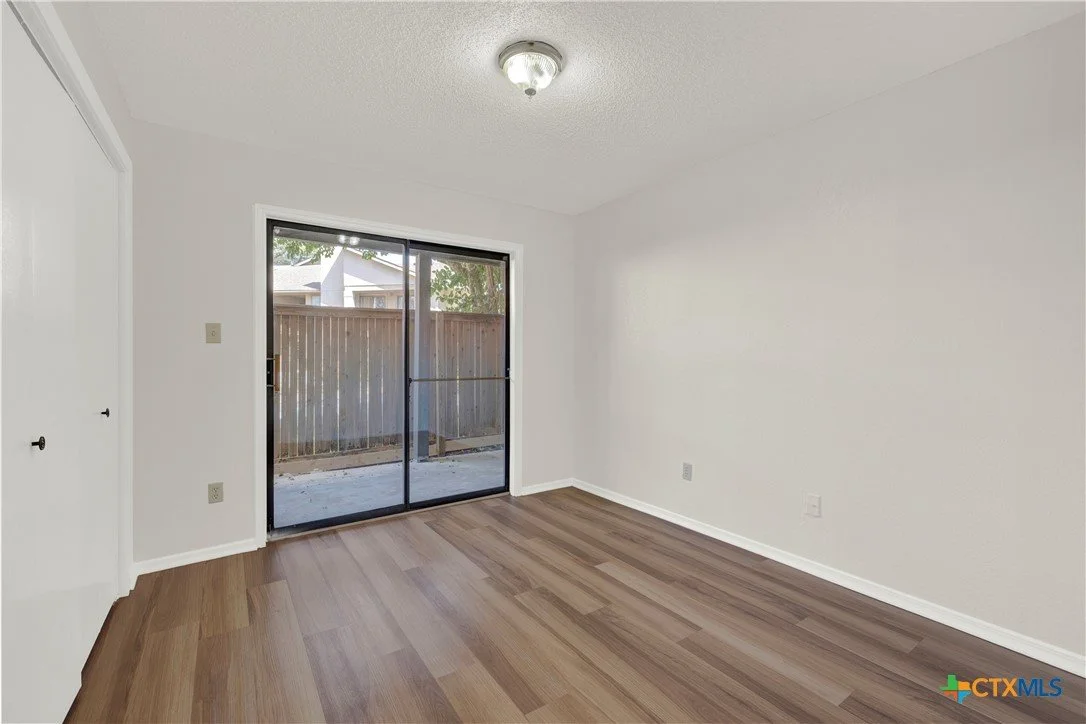 Empty room with hardwood floors, white walls, a sliding glass door leading to a patio, a ceiling light fixture, and a closet with white doors.