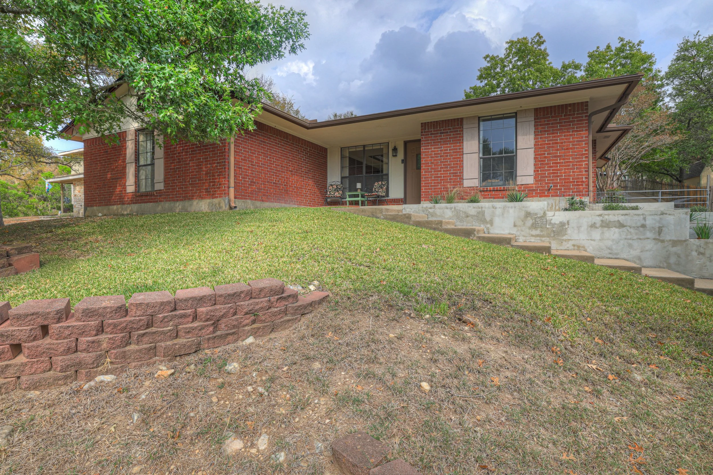 A brick house with a sloped yard, concrete stairs, and a front porch with two chairs, surrounded by green trees and cloudy sky.