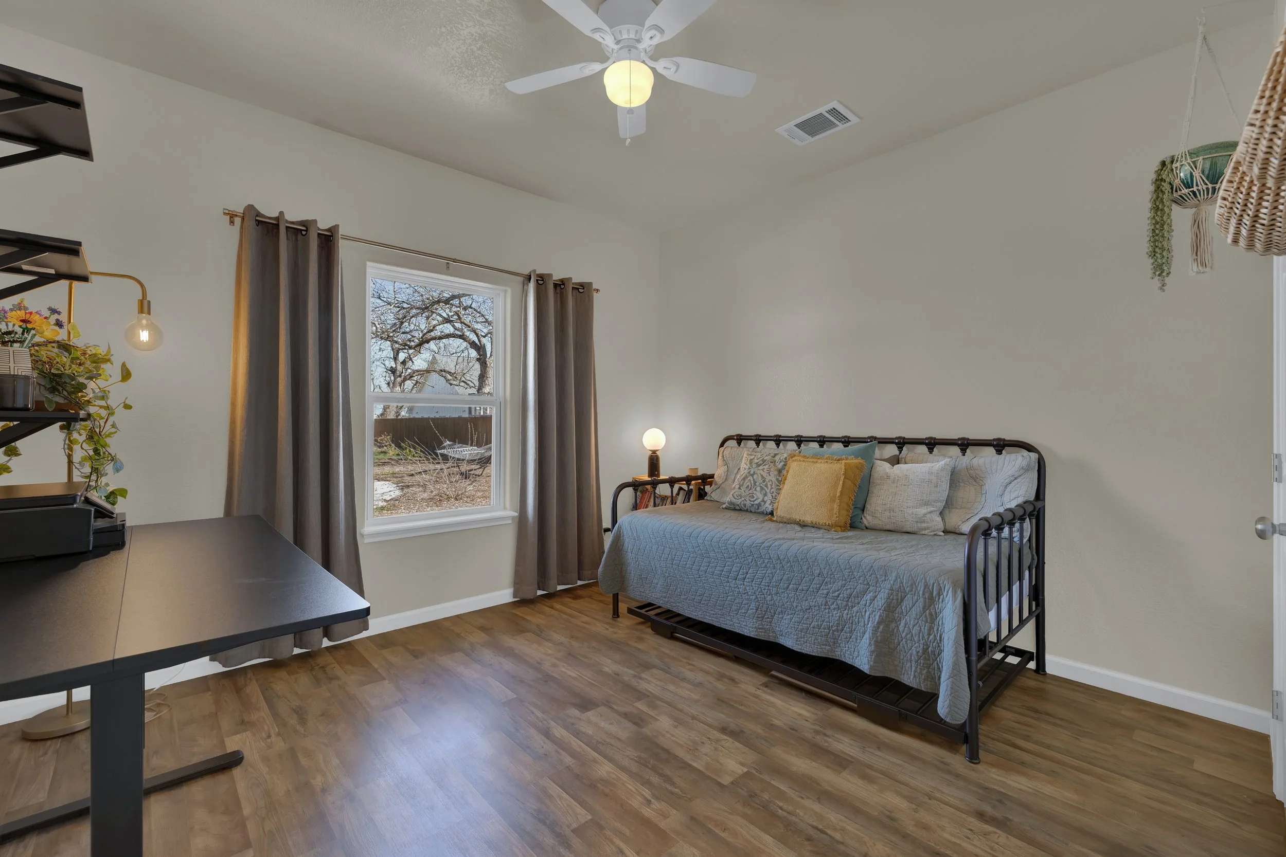 A bedroom with white walls, hardwood flooring, a window with brown curtains, a black daybed with pillows, a black desk, and a ceiling fan with a light fixture.