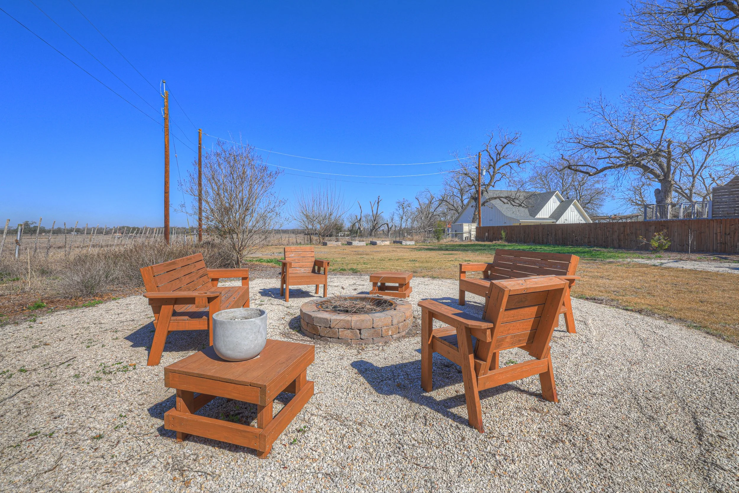 Outdoor backyard scene with five wooden chairs arranged around a brick fire pit on gravel, a white concrete pot on a small wooden table, dry trees, a grassy lawn, a wooden fence, power lines, and a house with a white roof under a clear blue sky.