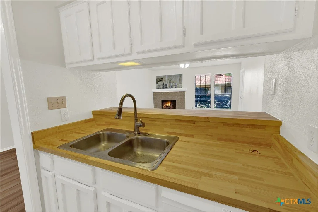 Kitchen with a wooden countertop, white cabinets, a double sink, and a view into a living room with a fireplace and windows.