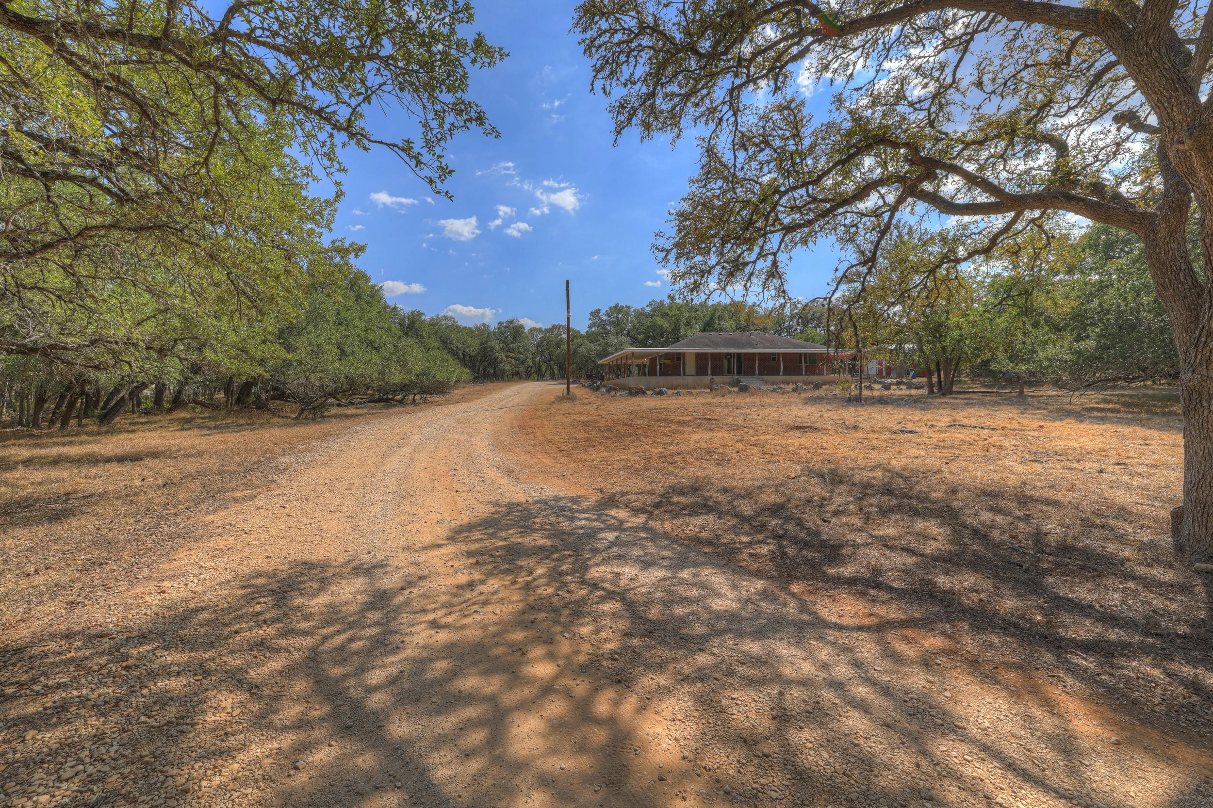 A rural dirt driveway leading to a house surrounded by trees, with a blue sky and scattered clouds overhead.