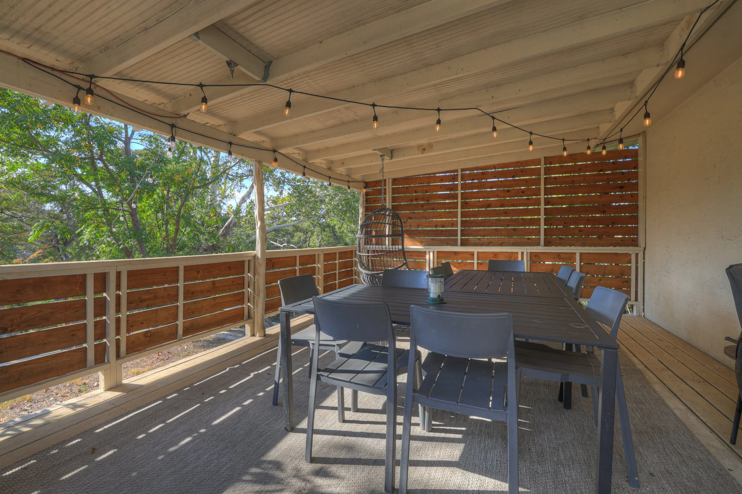 Covered outdoor patio with string lights, a large dining table, and a hanging egg chair, overlooking green trees.