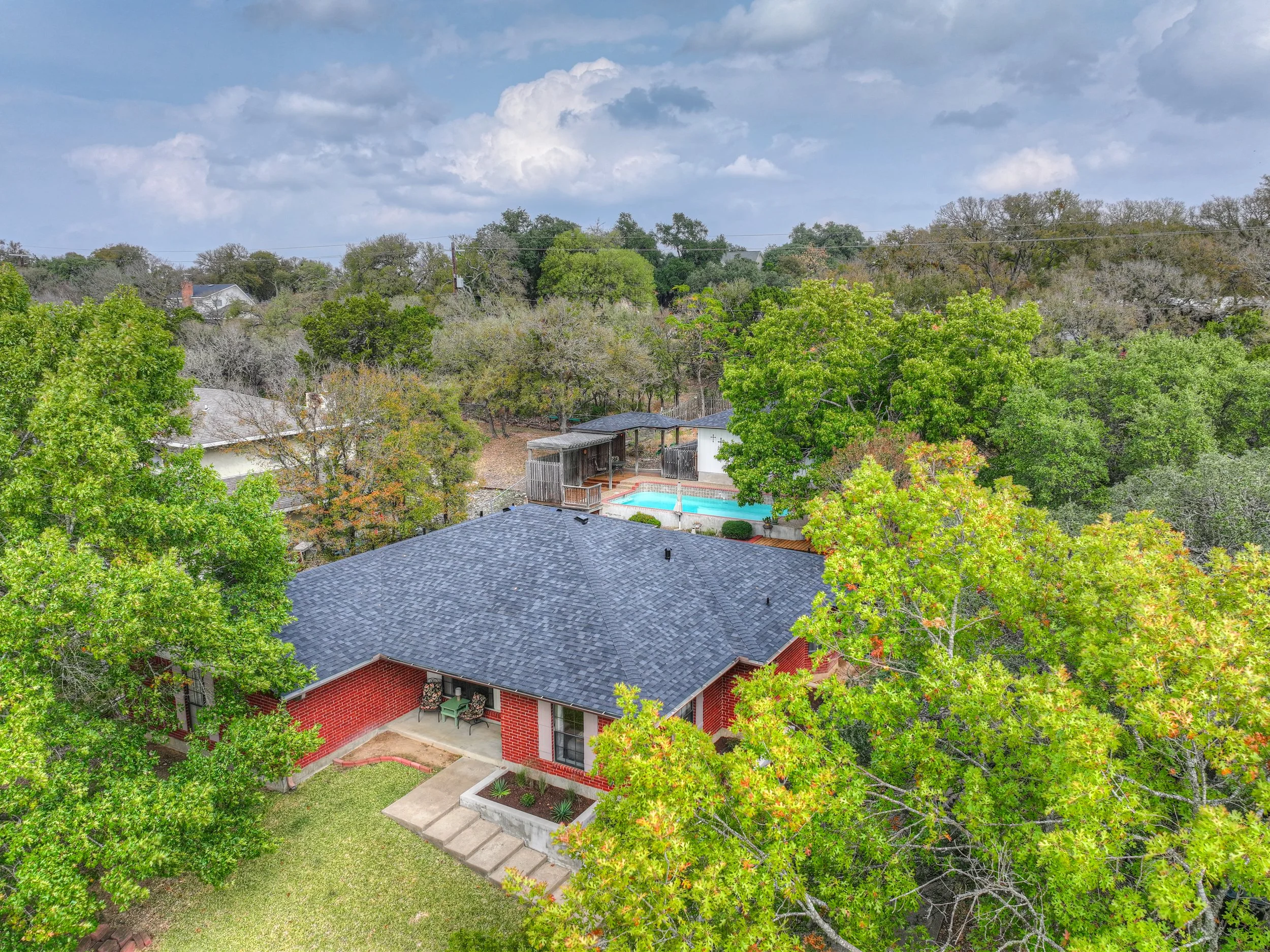 Aerial view of a suburban home with a dark gray roof, brick exterior, and a small front porch with seating. The backyard features a swimming pool and hot tub, surrounded by a wooden deck and various trees with green foliage. The sky is partly cloudy 