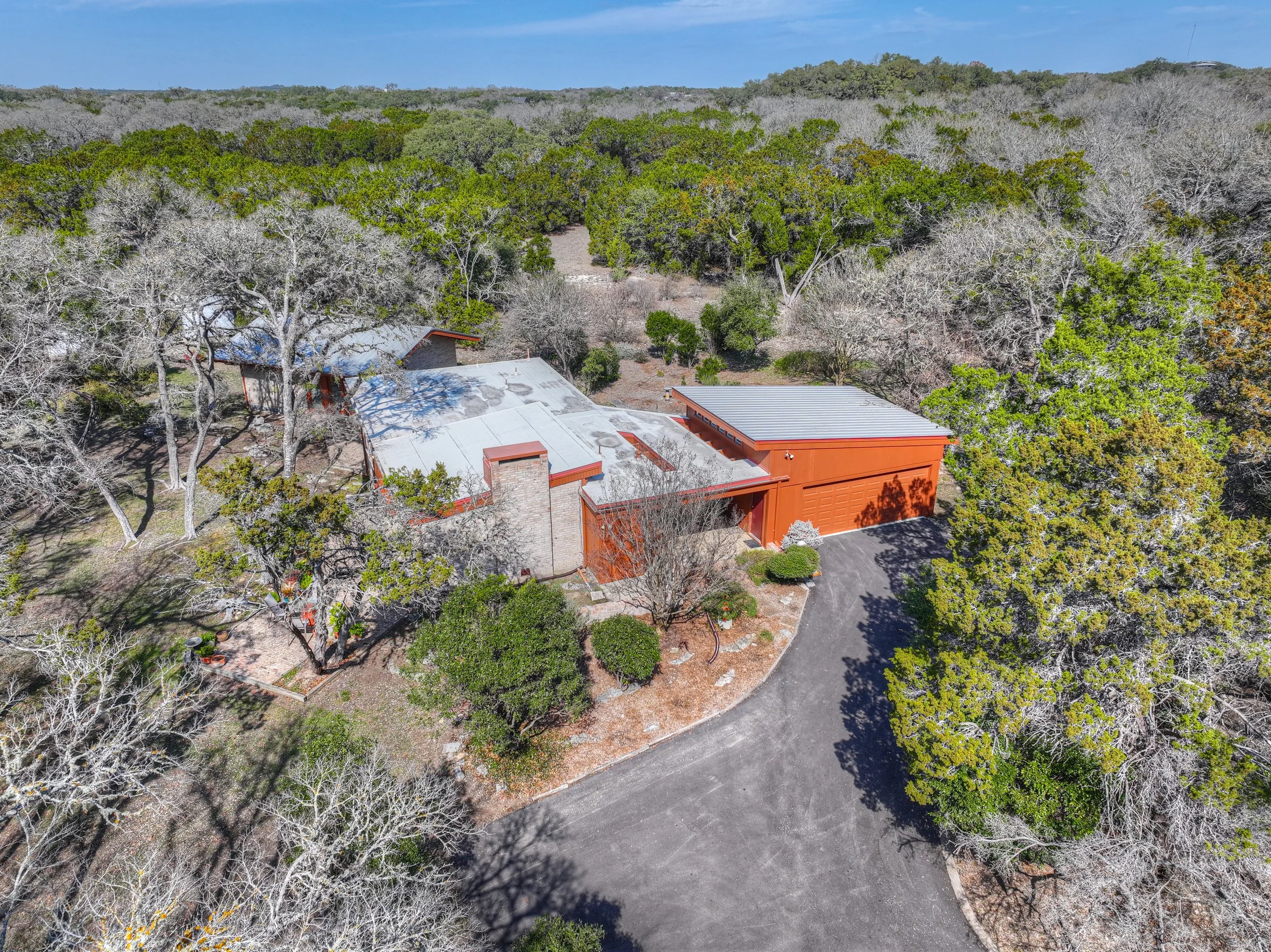 Aerial view of a house surrounded by trees with a mix of green and leafless branches, a curved driveway leading to a garage, and a forested landscape in the background.