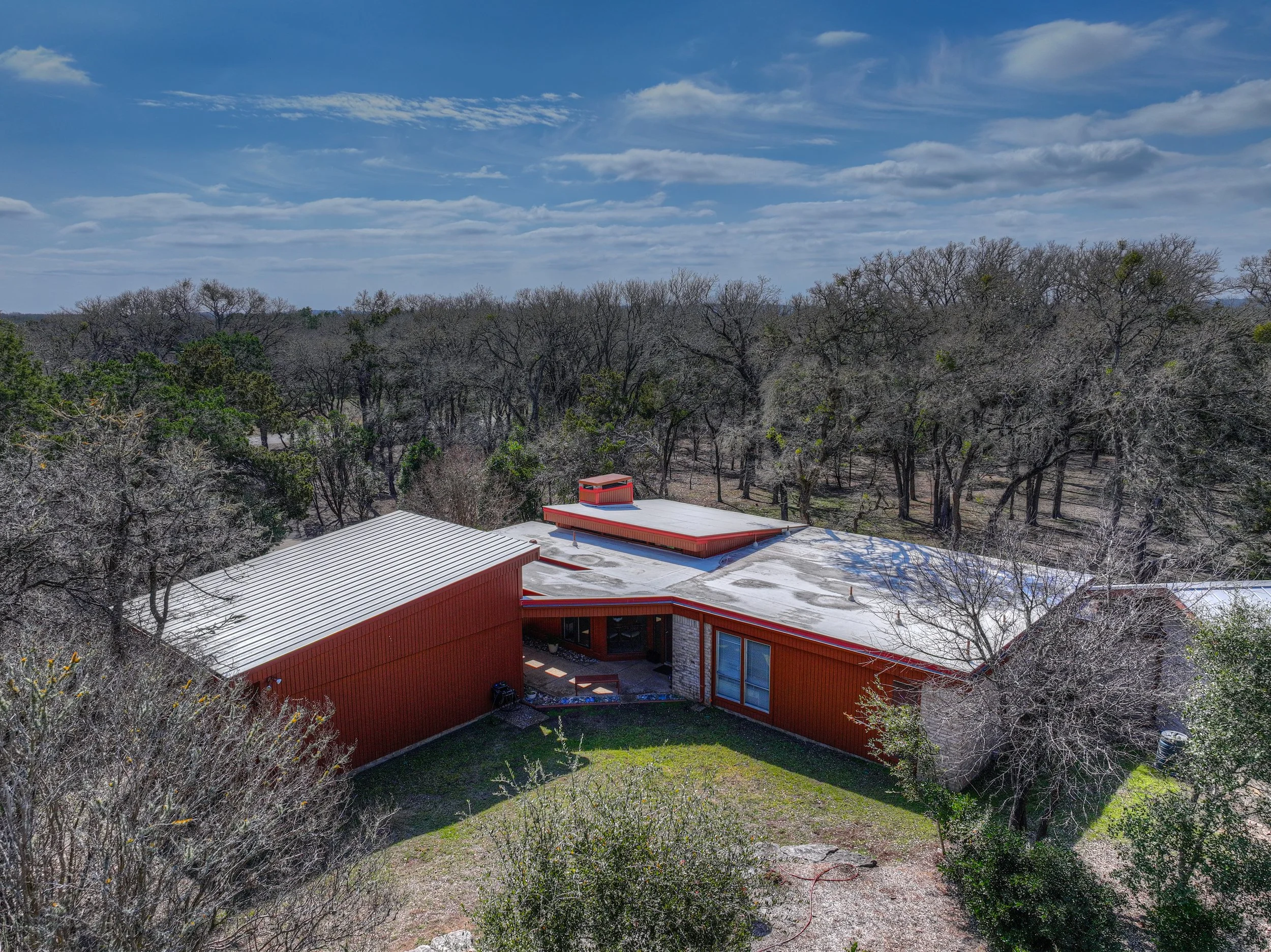 Aerial view of a modern red building with a white roof, surrounded by leafless trees and a clear blue sky with scattered clouds.