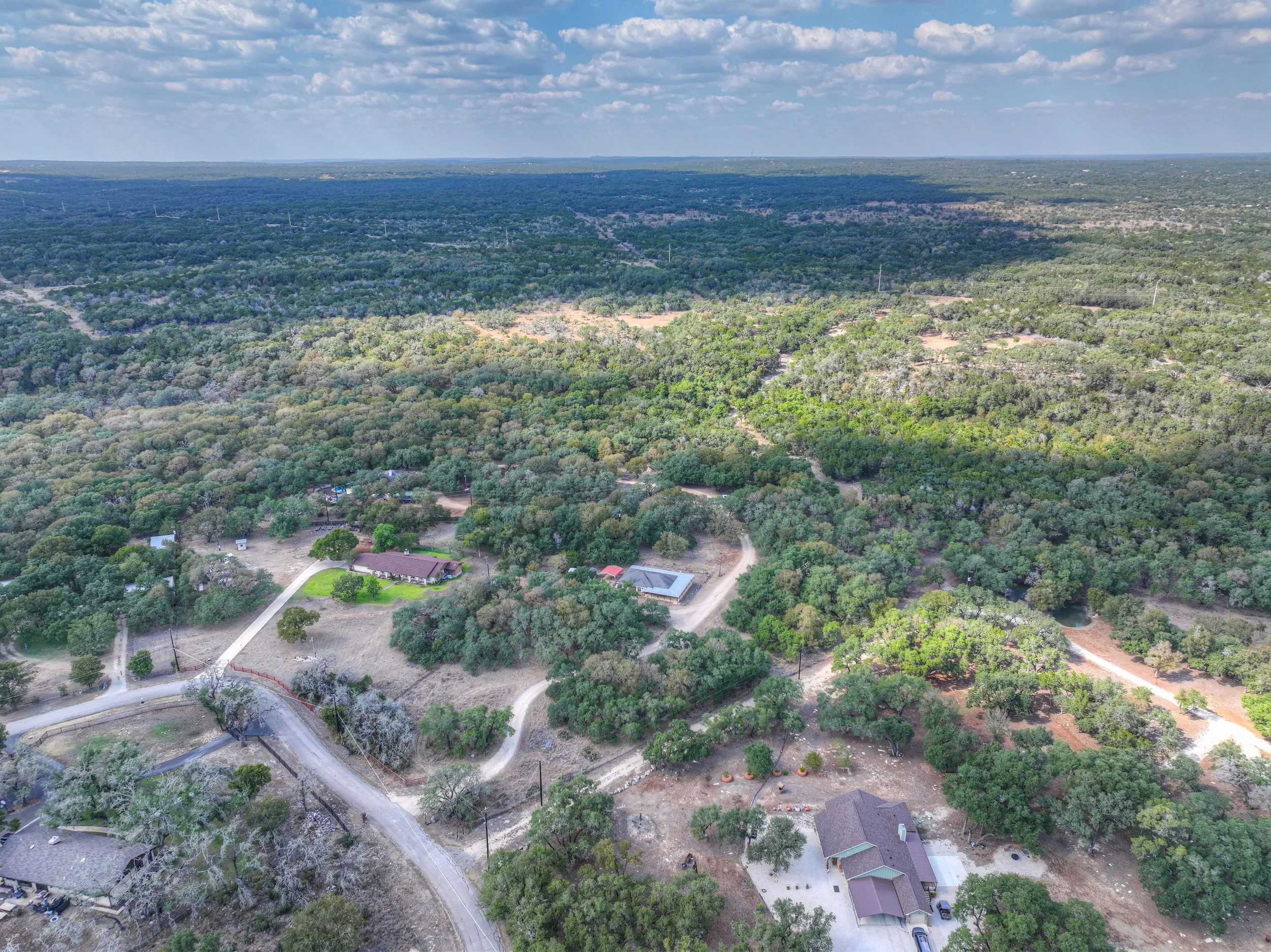 Aerial view of a rural area with trees, houses, winding roads, and open land under a partly cloudy sky.