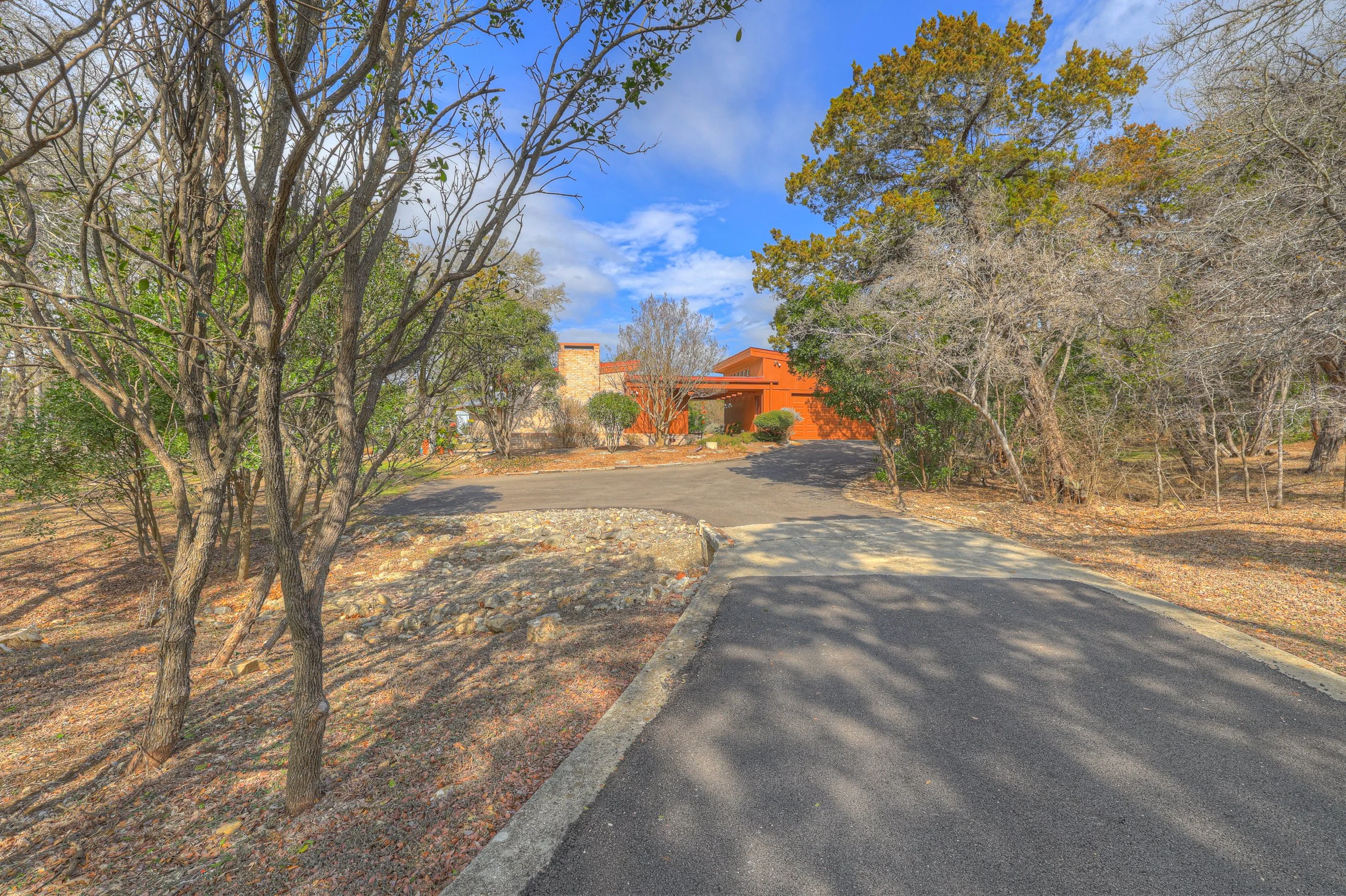 A winding paved driveway leading to a brick house with orange exterior walls, surrounded by trees with some leaves on the ground and some trees bare, under a partly cloudy blue sky.