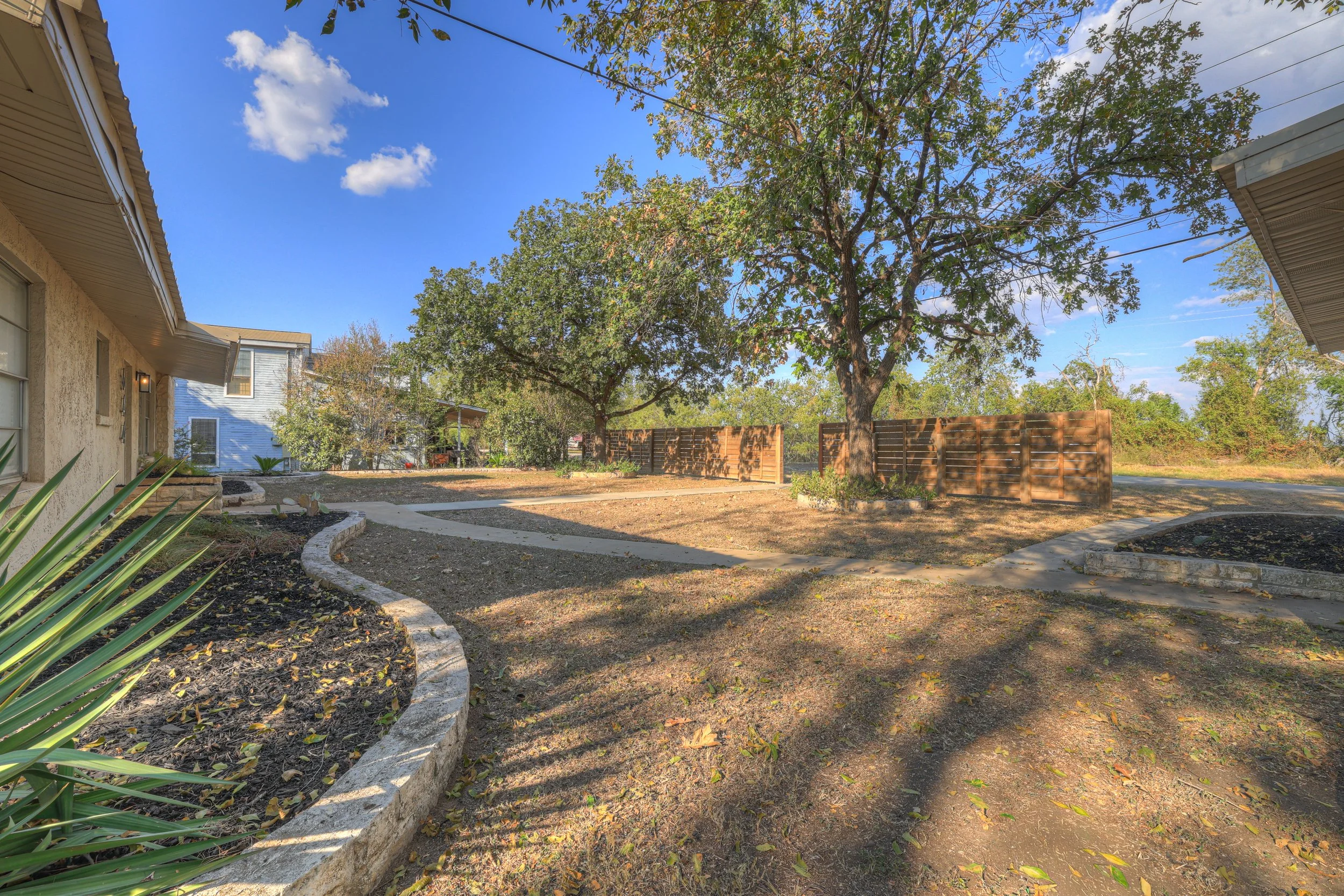 View of a residential backyard with trees, a curved path, a newly planted garden bed with a stone border, and wooden privacy fences, under a clear blue sky.