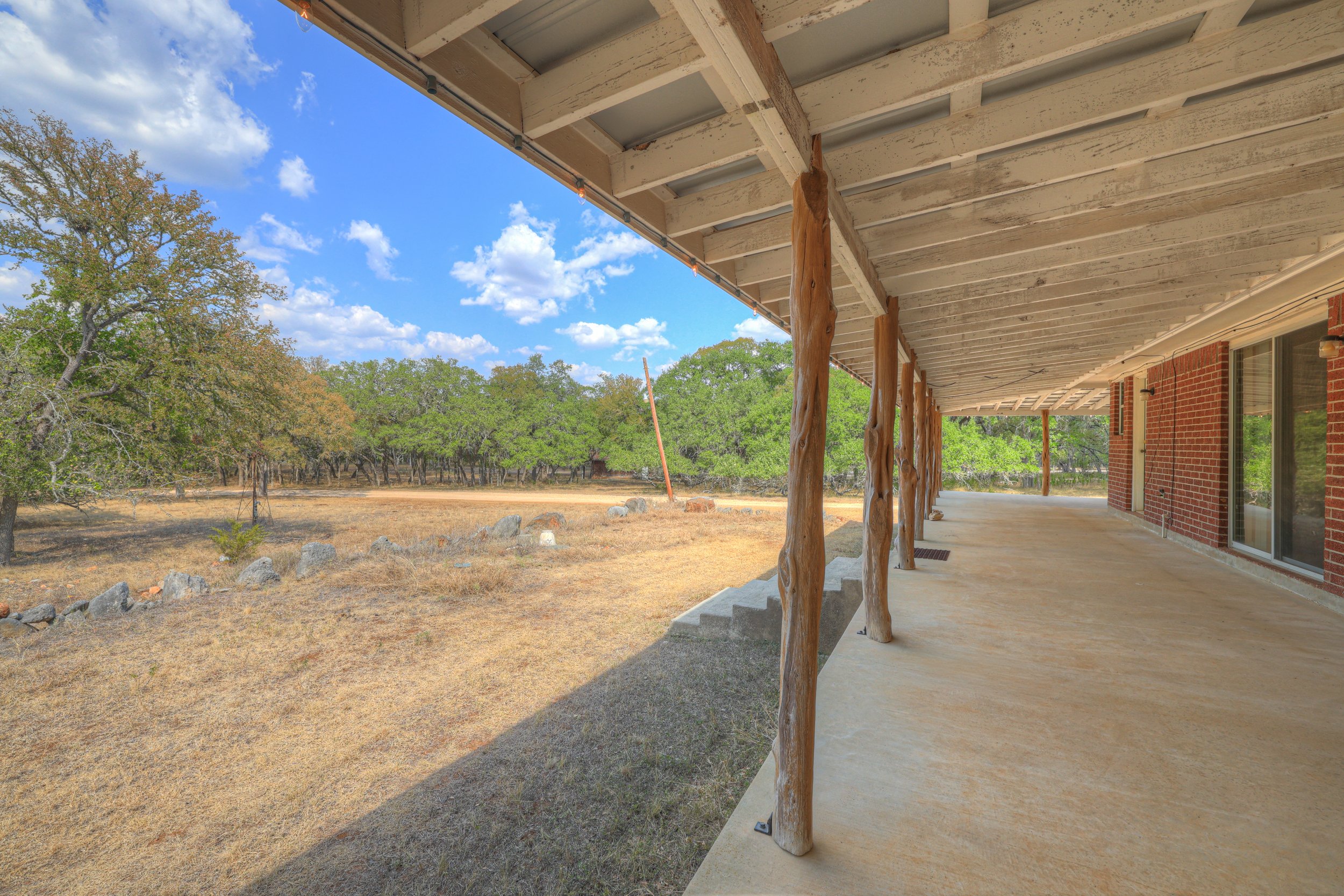 A covered patio with wooden support poles, overlooking a dry yard with trees and a blue sky with clouds.