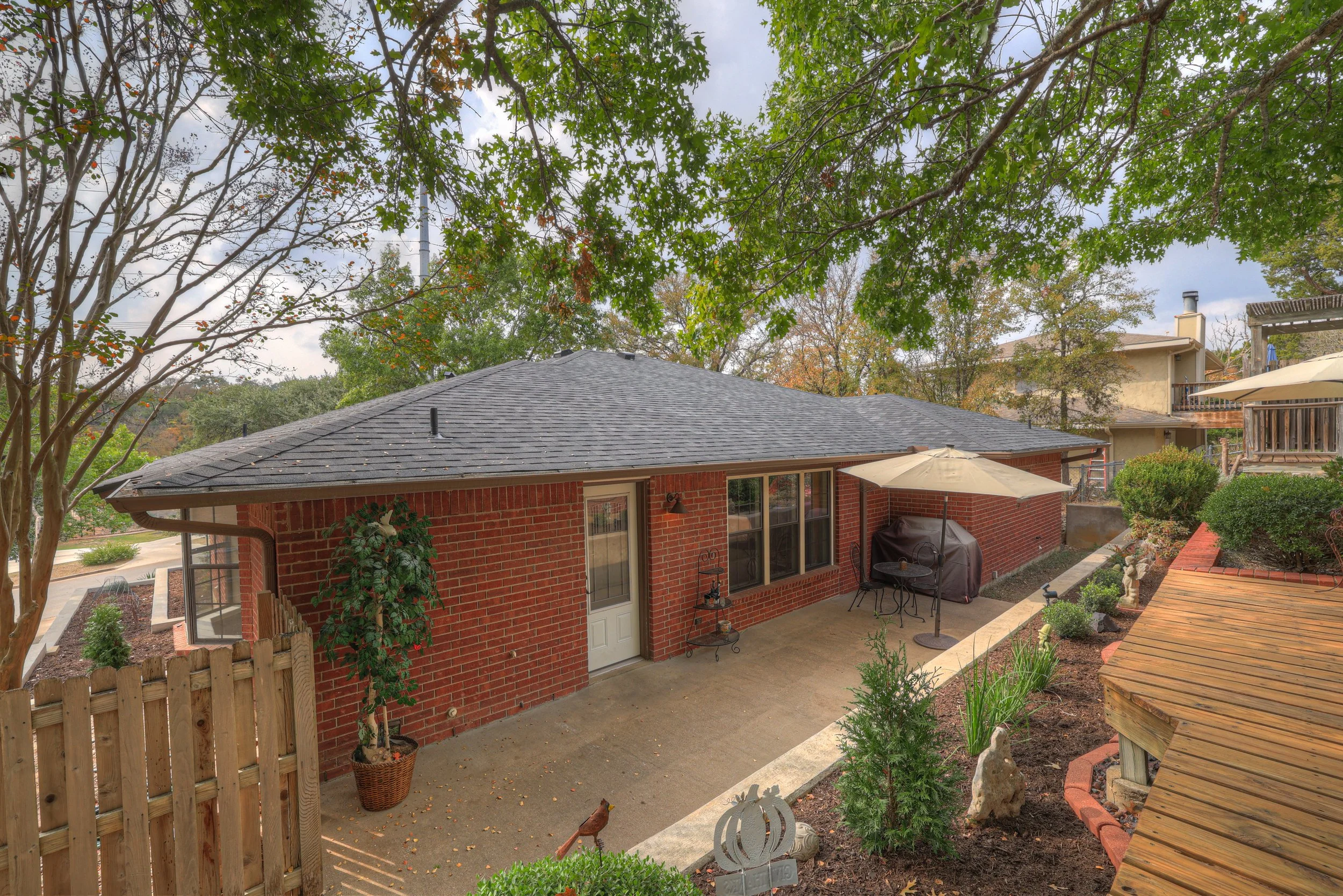 Backyard patio view of a brick house with a gray shingle roof, patio furniture with a large umbrella, potted plants, a covered grill, and surrounding greenery, trees, and neighboring houses.