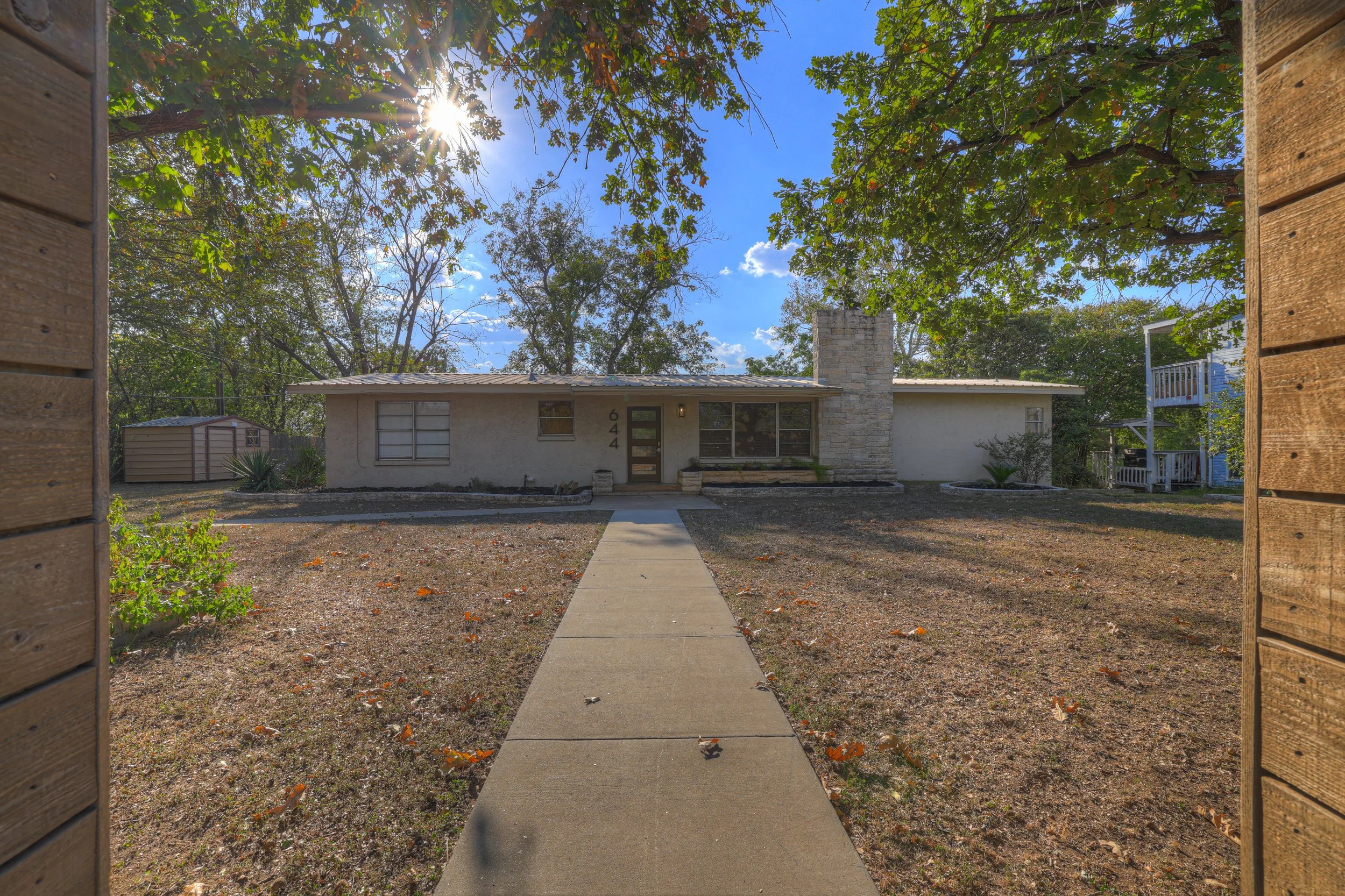 Front view of a single-story house with a sidewalk leading to the front door, surrounded by trees with sunlight filtering through the leaves, a small lawn area, and a shed in the yard.