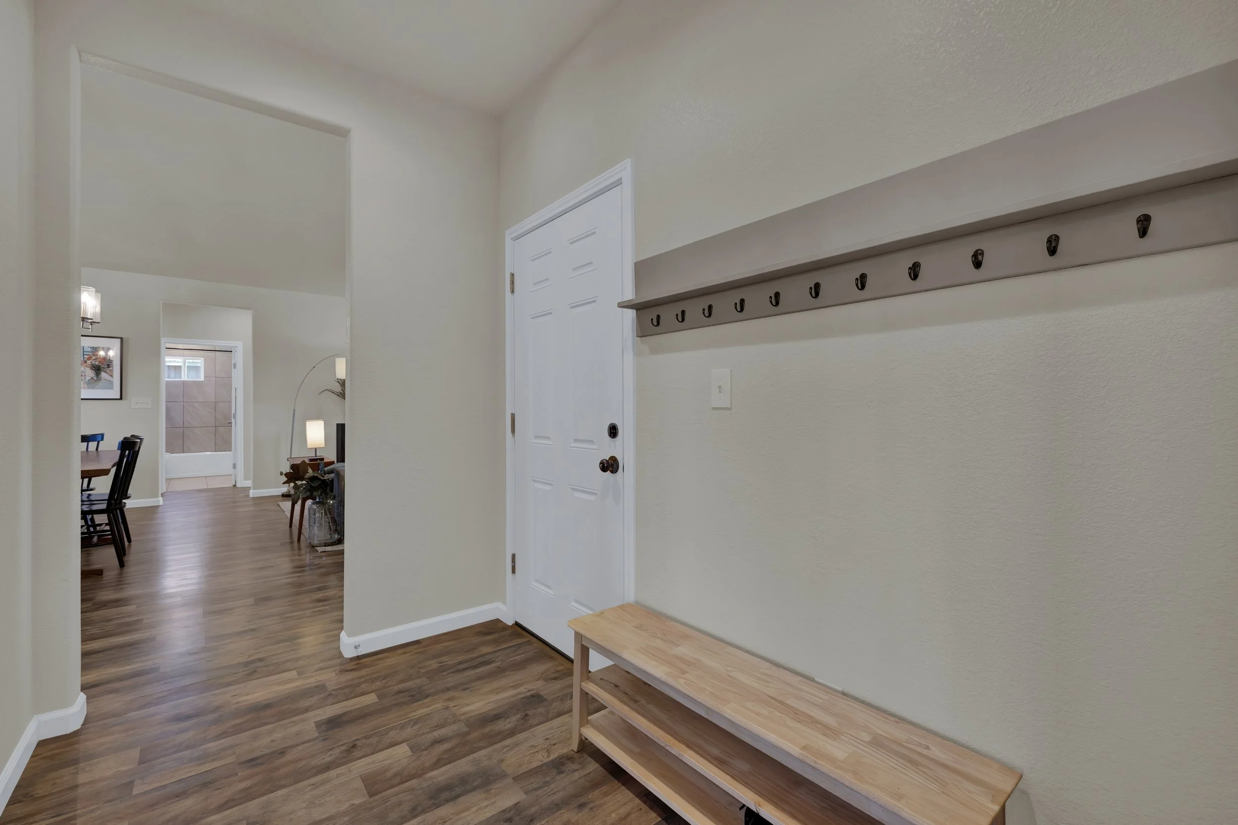 Empty entryway with a wooden bench, a wall-mounted coat rack, and a door leading into a living room with wood flooring and furniture.