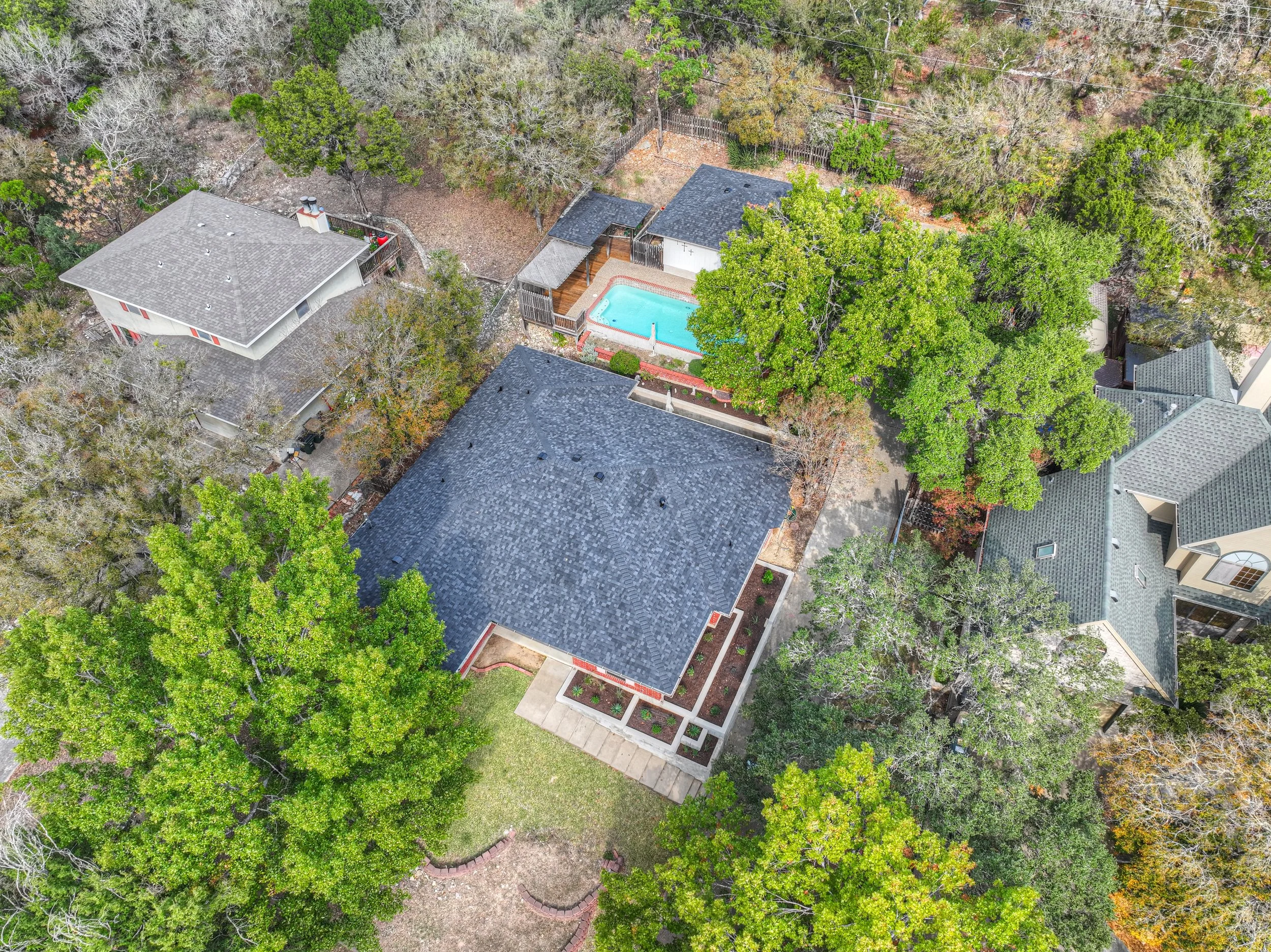 An aerial view of a residential backyard with a swimming pool, surrounded by trees, fences, and neighboring houses.