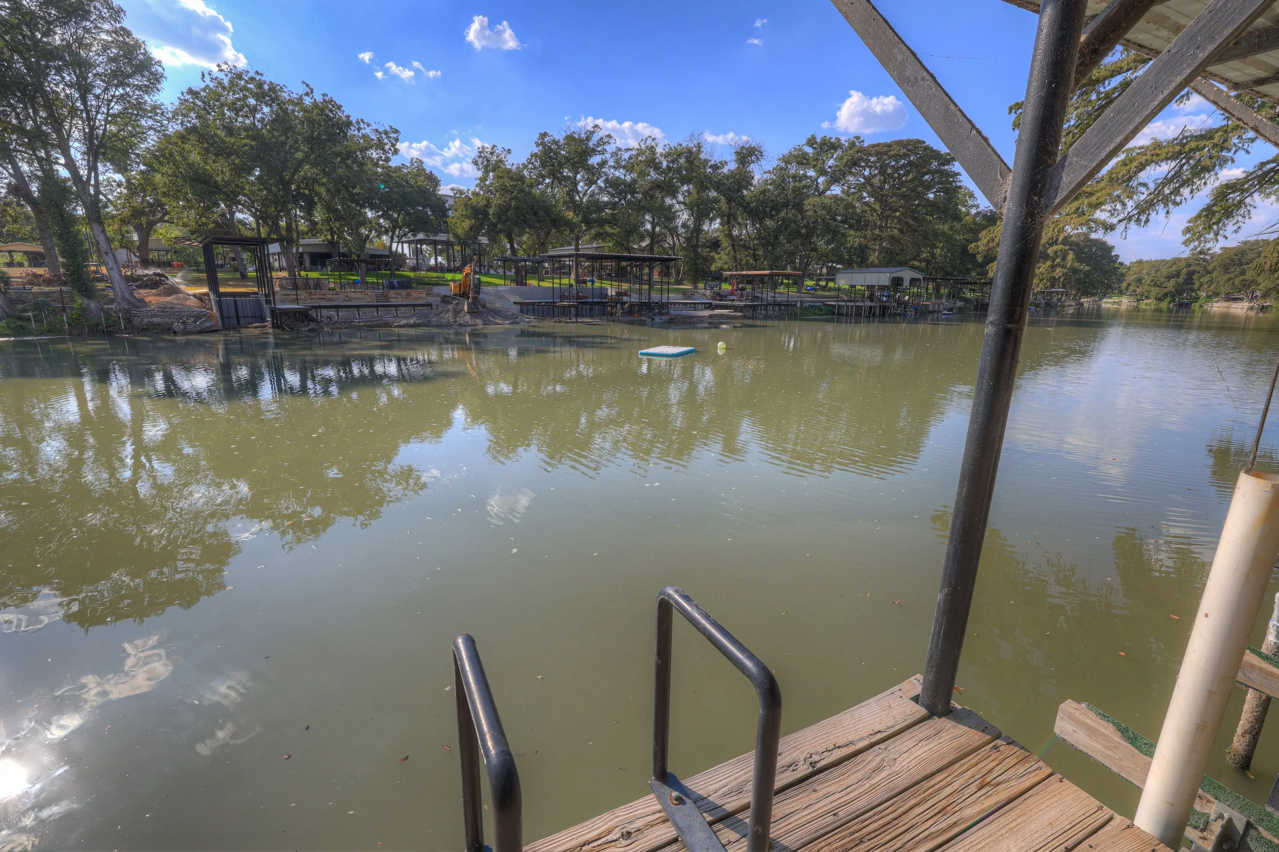 View of a waterfront with docks, trees, and a cloudy sky, as seen from a wooden dock with metal handrails.