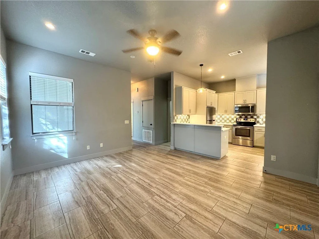 Open floor plan living room with gray walls, light wood flooring, large window with blinds, ceiling fan, and view of kitchen with white cabinets, stainless steel appliances, patterned backsplash, and island with a hanging light.