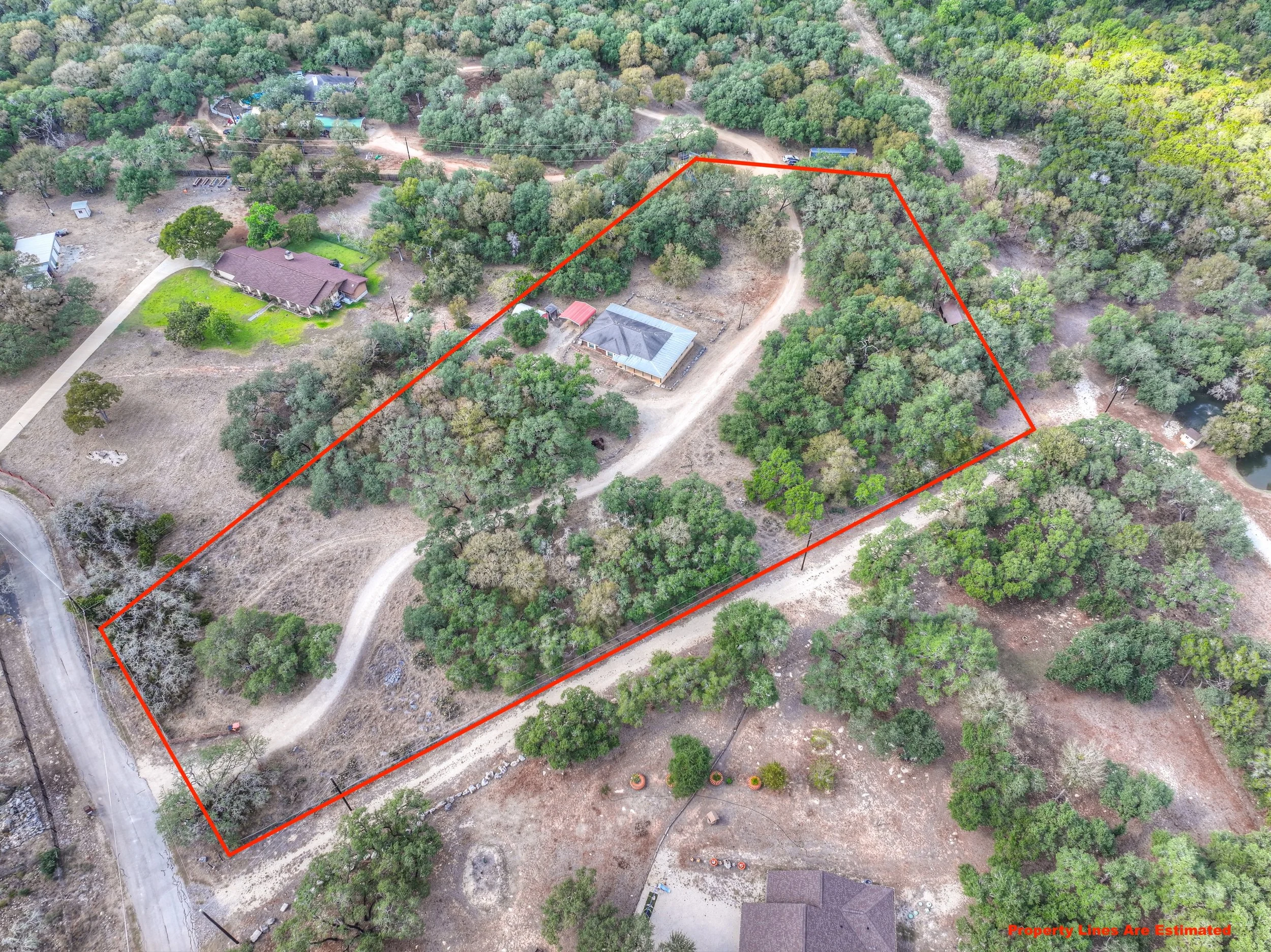 Aerial view of a large, wooded residential property outlined with a red property line. The property contains a house with a gray roof, a dirt driveway, and several trees, and is surrounded by other homes, trees, and natural landscape.