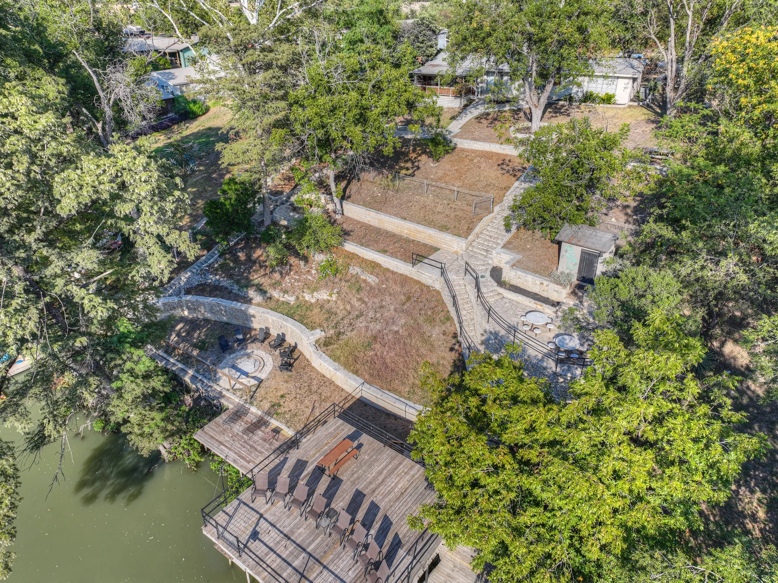 Aerial view of a backyard with outdoor seating, trees, a dock extending over water, and a staircase leading up to a garden area with a small shed.