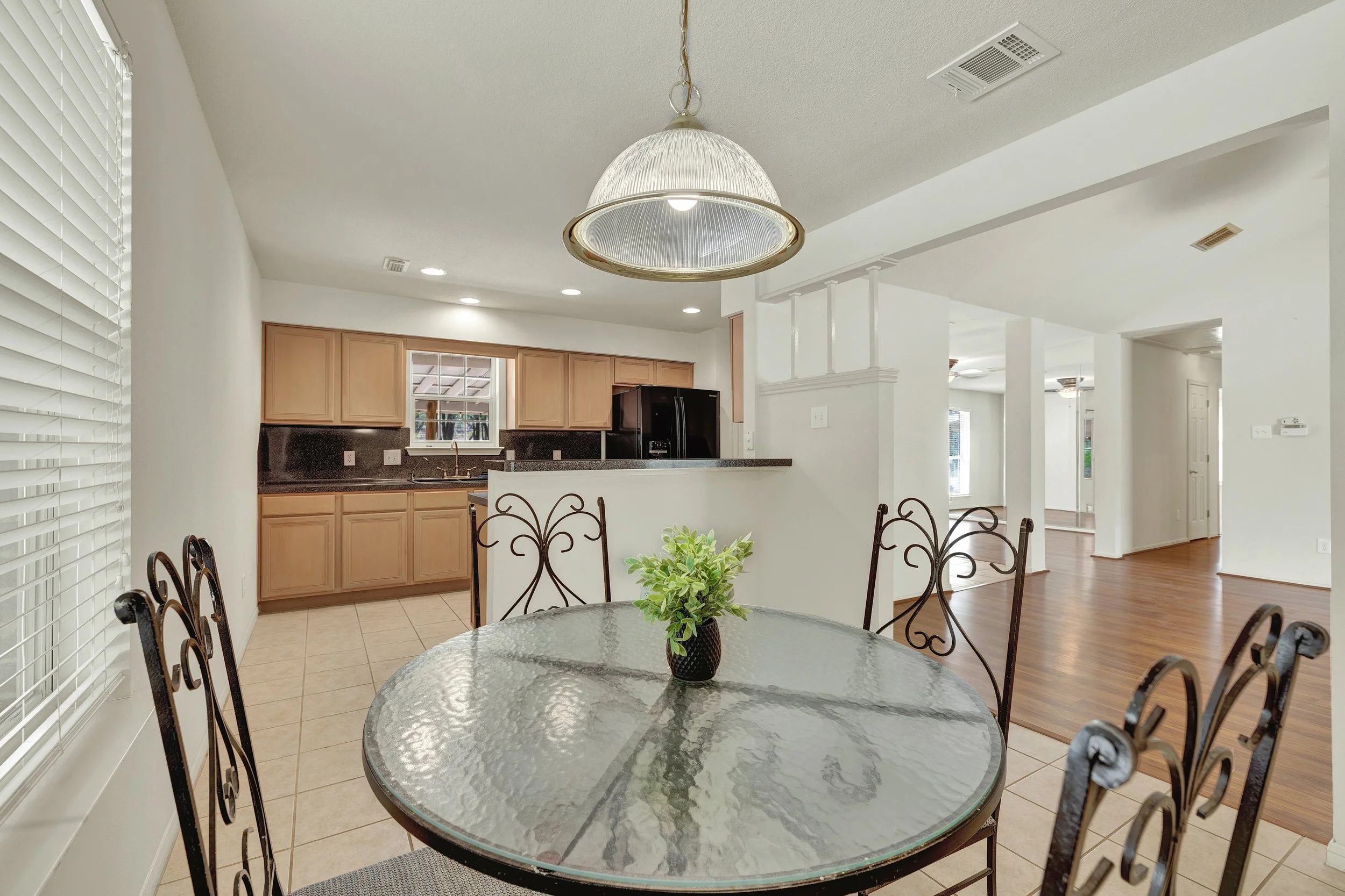 Dining area with glass table, metal chairs, and a potted plant centerpiece, adjacent to a kitchen with wooden cabinets and black refrigerator, in a bright, open-concept home.