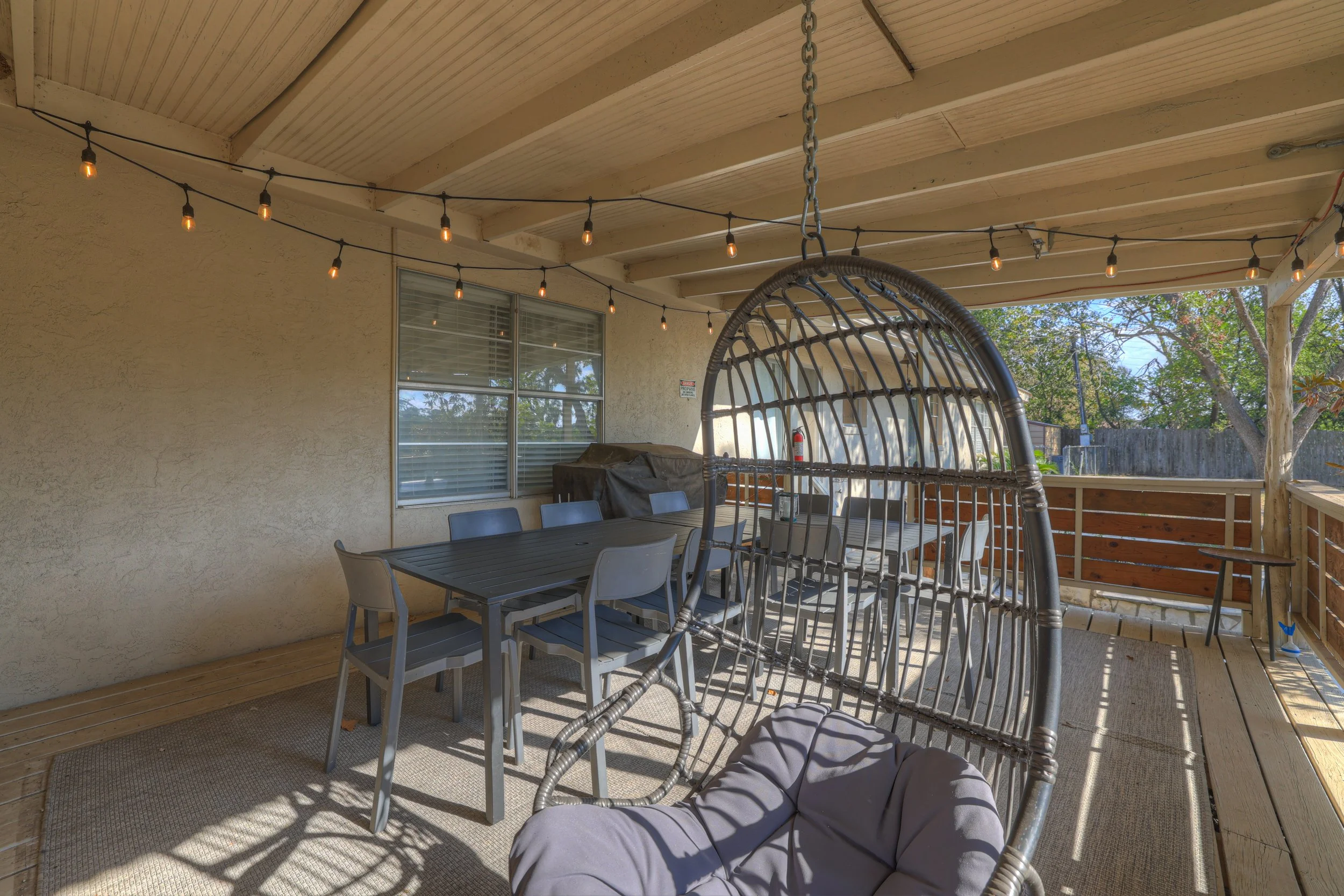Covered patio with outdoor dining table and chairs, hanging string lights, outdoor swing chair, and a privacy fence overlooking trees and a backyard.