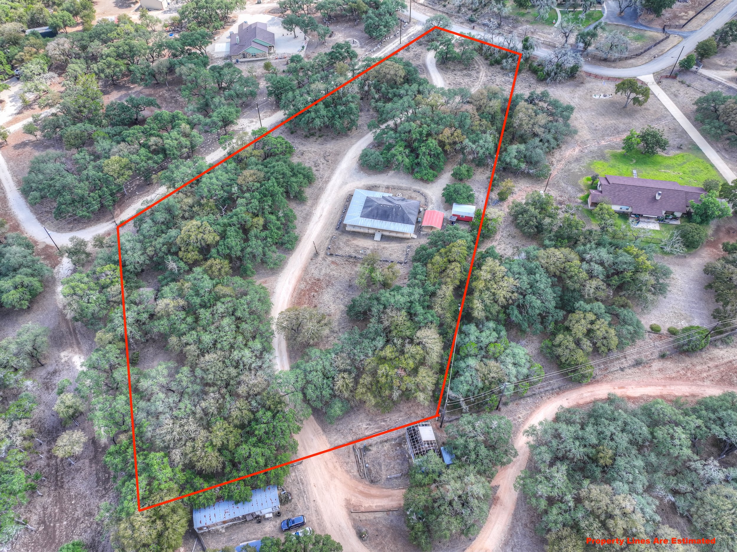 Aerial view of a large wooded property outlined with red property lines, featuring a house with a gray roof, a red shed, and a dirt driveway surrounded by trees.