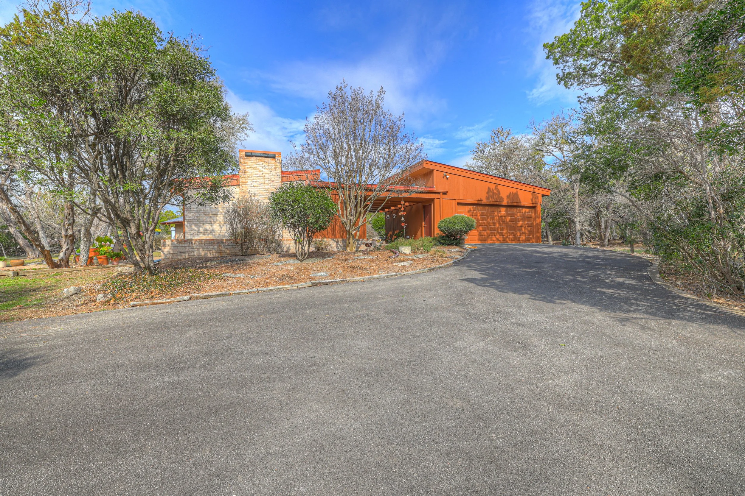 Front view of a mid-century modern house with a sloped roof, a brick chimney, and a driveway, surrounded by trees and shrubs on a clear day.