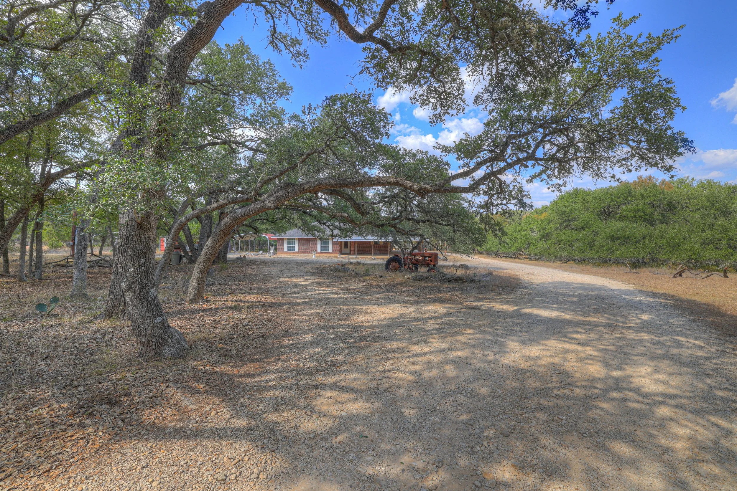 A gravel driveway lined with large trees leading to a brick house in the background, with a vintage tractor parked near the trees.