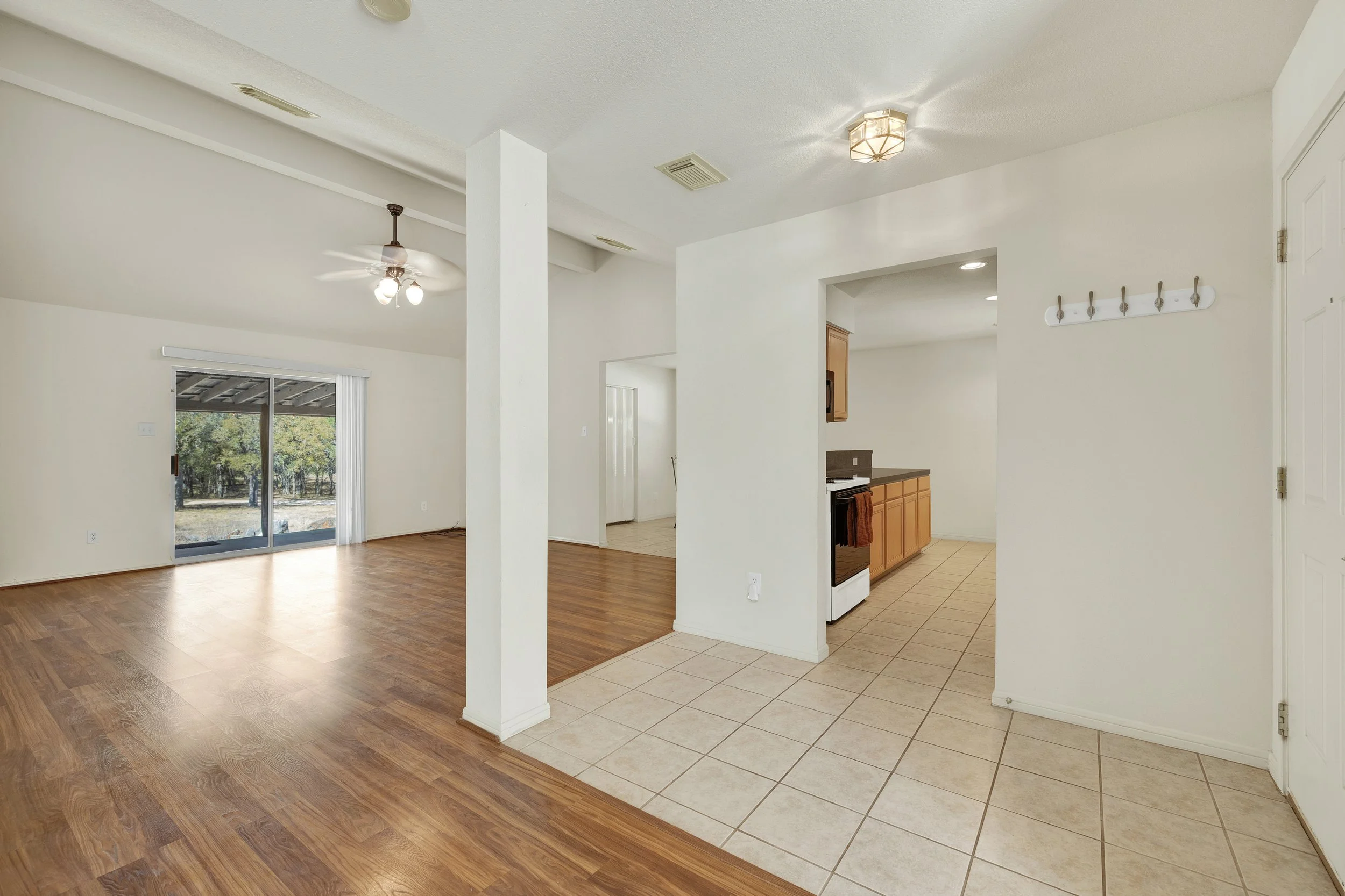 Living room with wood flooring, sliding glass door to outside, ceiling fan, and white walls. Kitchen area with tile flooring, wooden cabinets, and a stove.