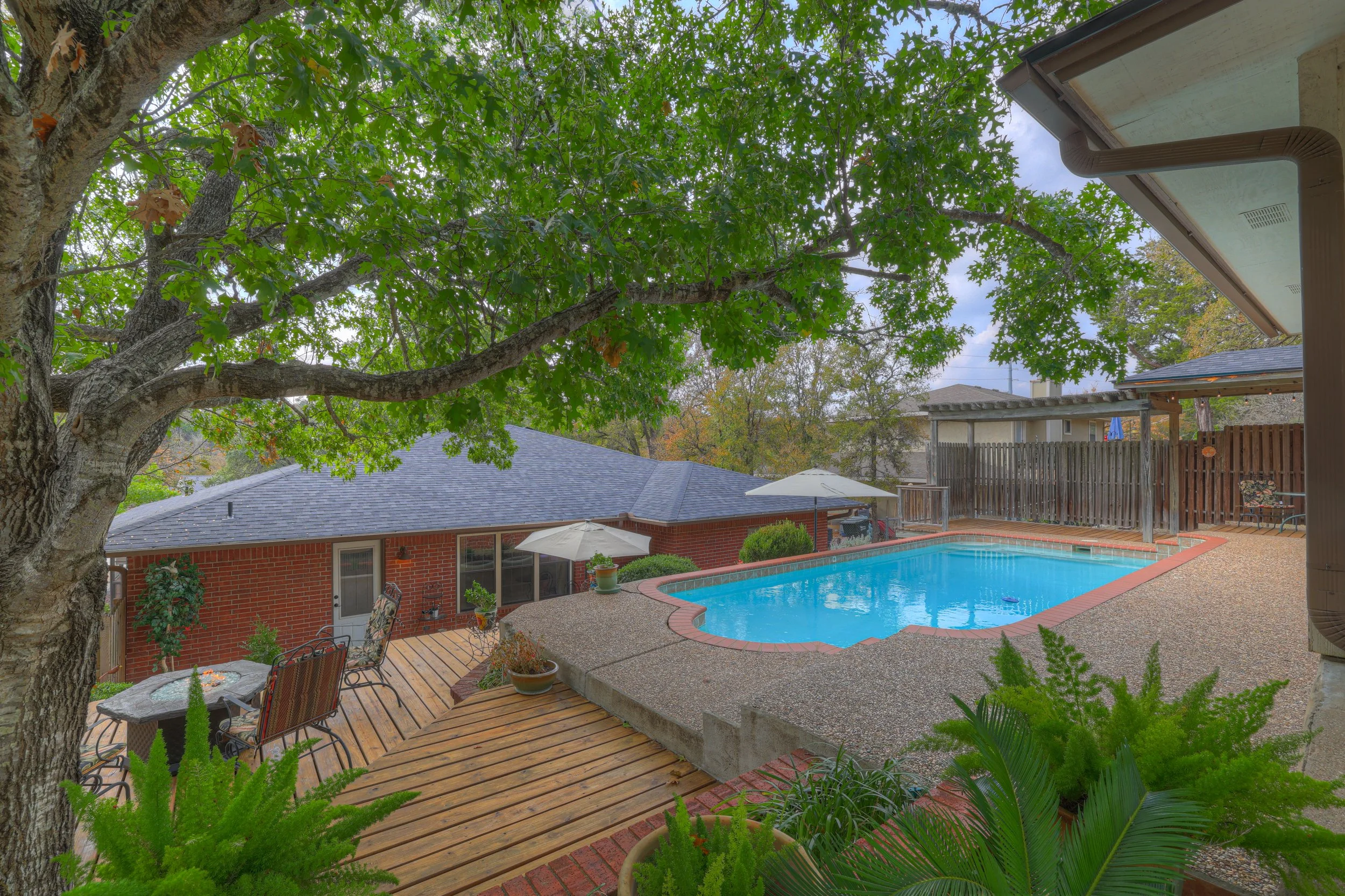 A backyard view featuring a swimming pool surrounded by a concrete deck, a wooden deck area with outdoor furniture and umbrellas, a tree with green leaves, and a fence in the background. The house wall with a window and door is visible behind the poo