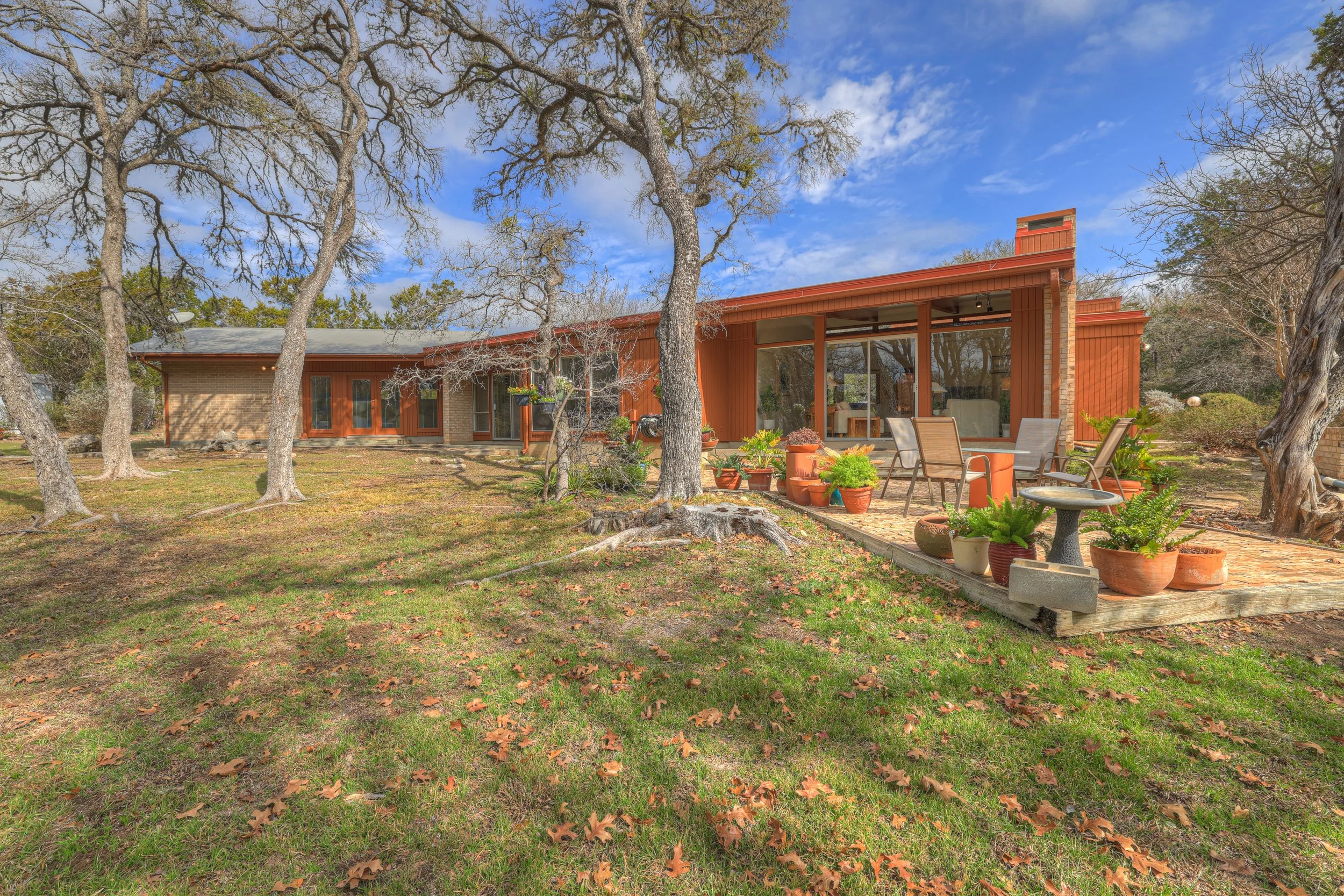A backyard with a deck featuring potted plants, patio chairs, a birdbath, trees with some leaves fallen, and a mid-century modern house with large windows under a partly cloudy sky.