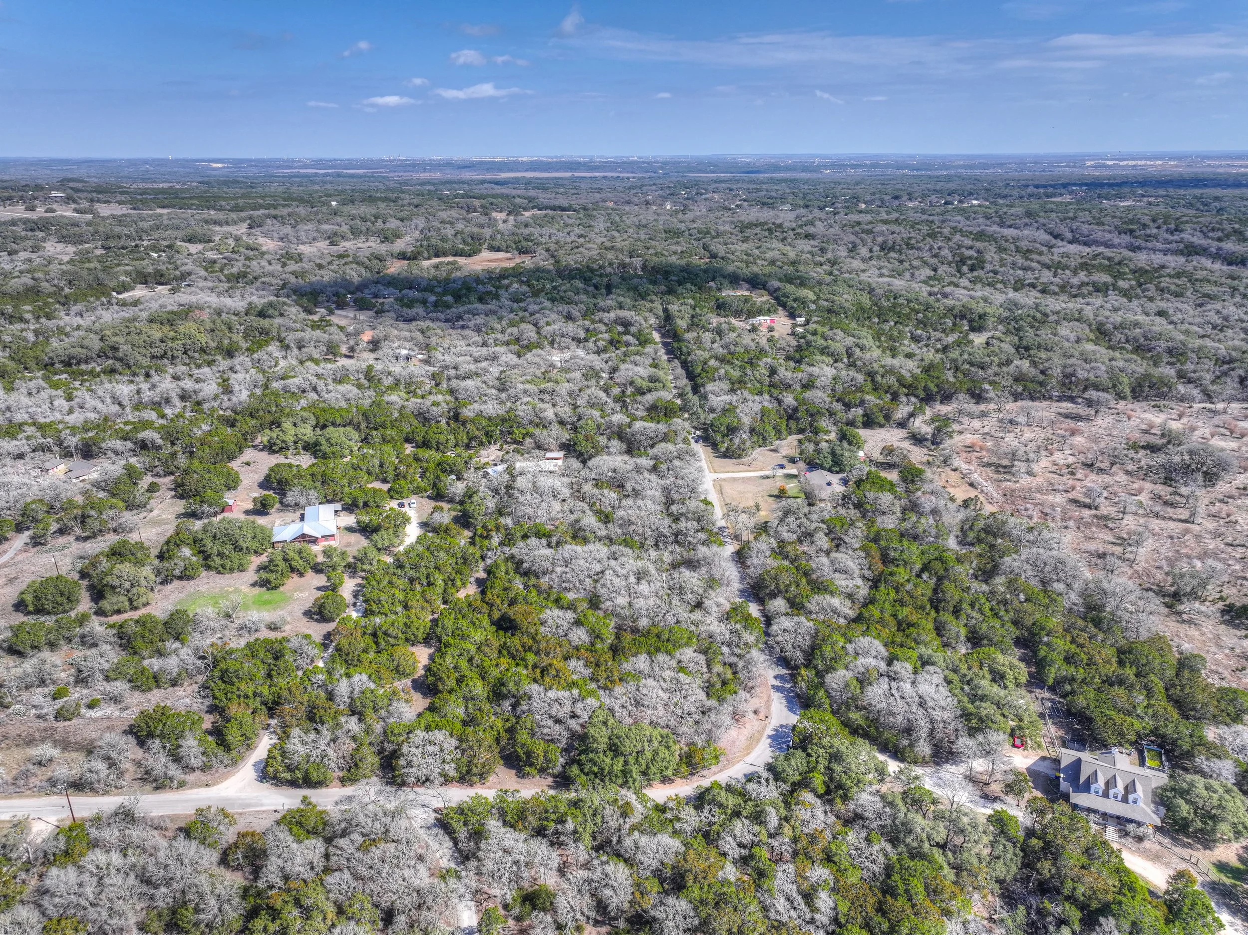 Aerial view of a rural area with dense trees, scattered houses, and winding roads under a partly cloudy sky.