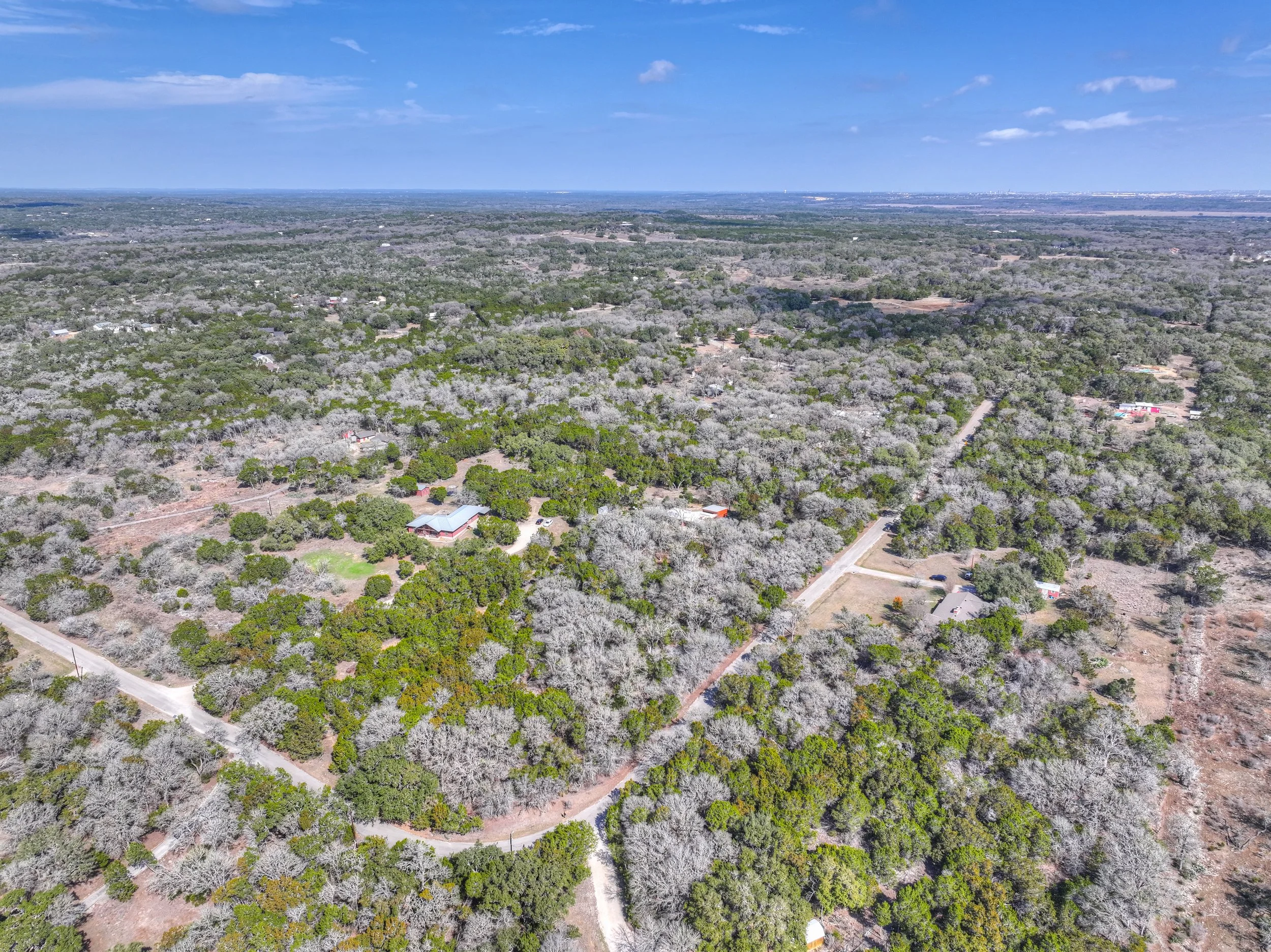 Aerial view of a rural landscape with scattered houses and trees, some with bare branches, under a partly cloudy sky.