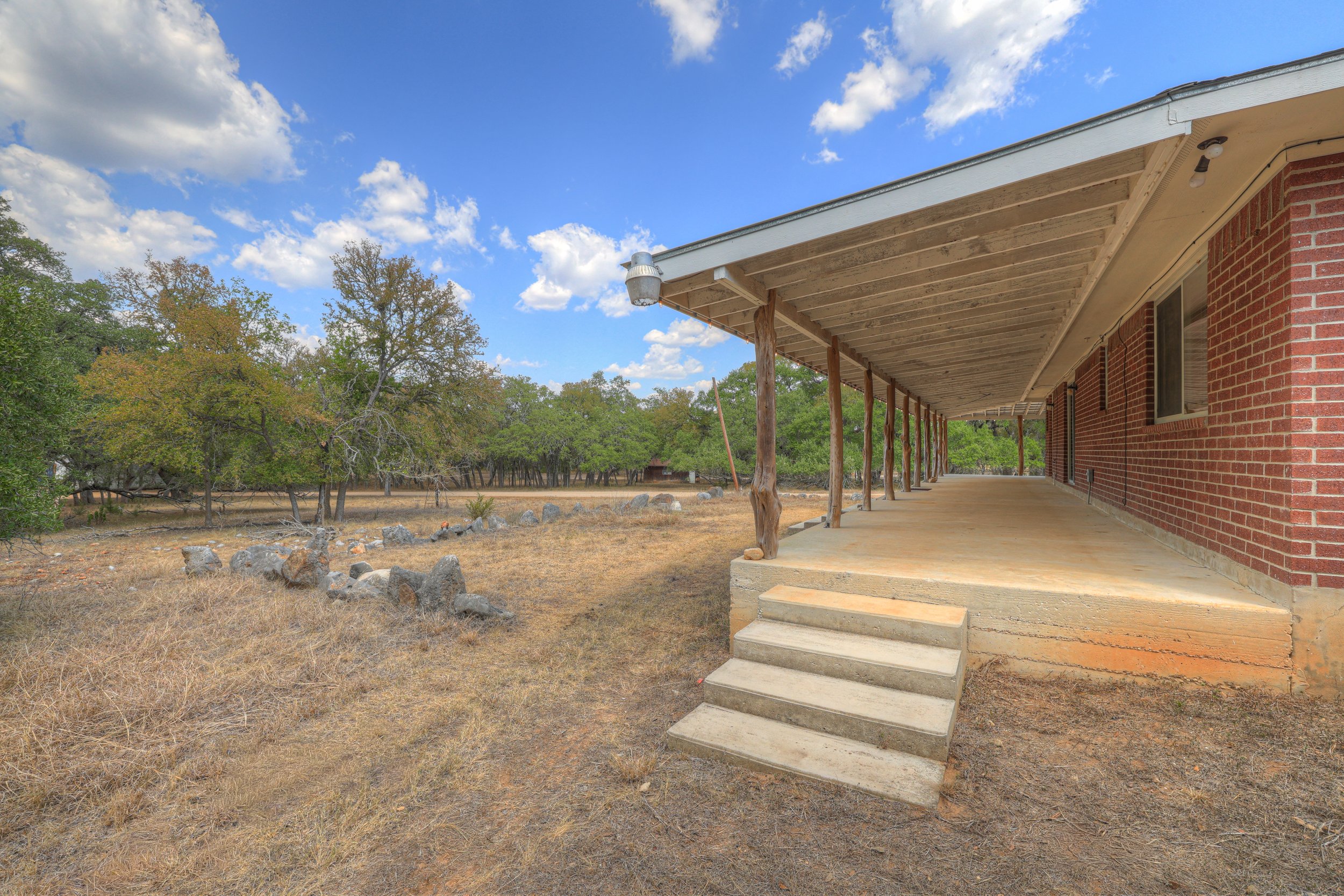 Front porch of a red brick house with wooden support beams, steps leading up to the porch, and a dry grassy yard with rocks and trees in the background under a partly cloudy sky.