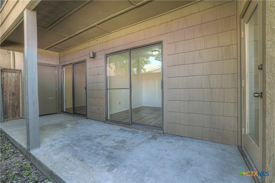 Image of a covered patio with sliding glass doors leading to an interior room. The patio floor is concrete, and the exterior wall has horizontal siding.