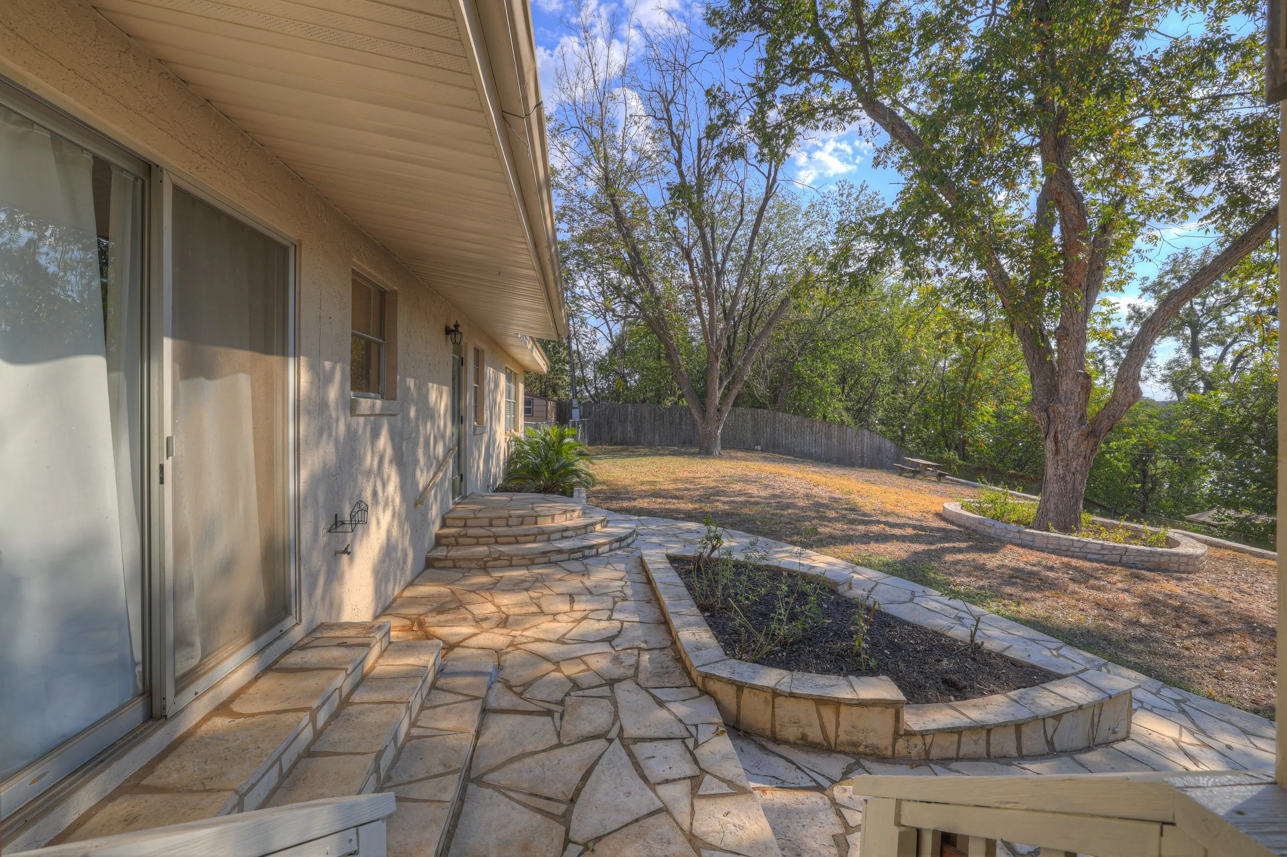 Backyard patio with stone pathway, small garden bed, large tree, wooden fence, and picnic table under a clear blue sky.