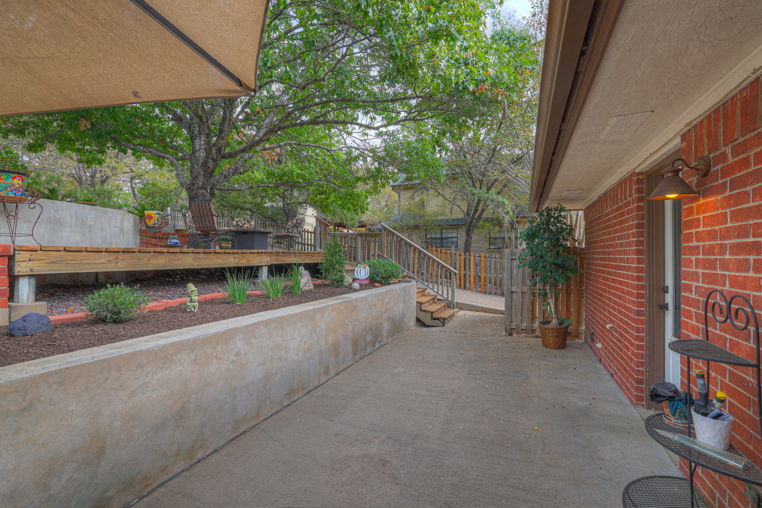 A backyard patio with a concrete floor, a brick house wall, a small potted tree, and a black metal shelf with containers, against the house wall. A wooden staircase leads up to a garden with a large tree, and a grassy yard with outdoor furniture and 