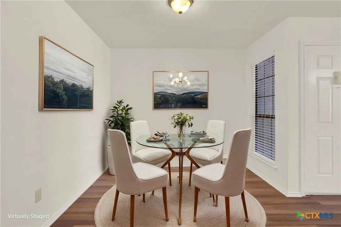 A dining room with a round glass table, four white upholstered chairs, a vase with flowers, and outdoor landscape paintings on white walls.