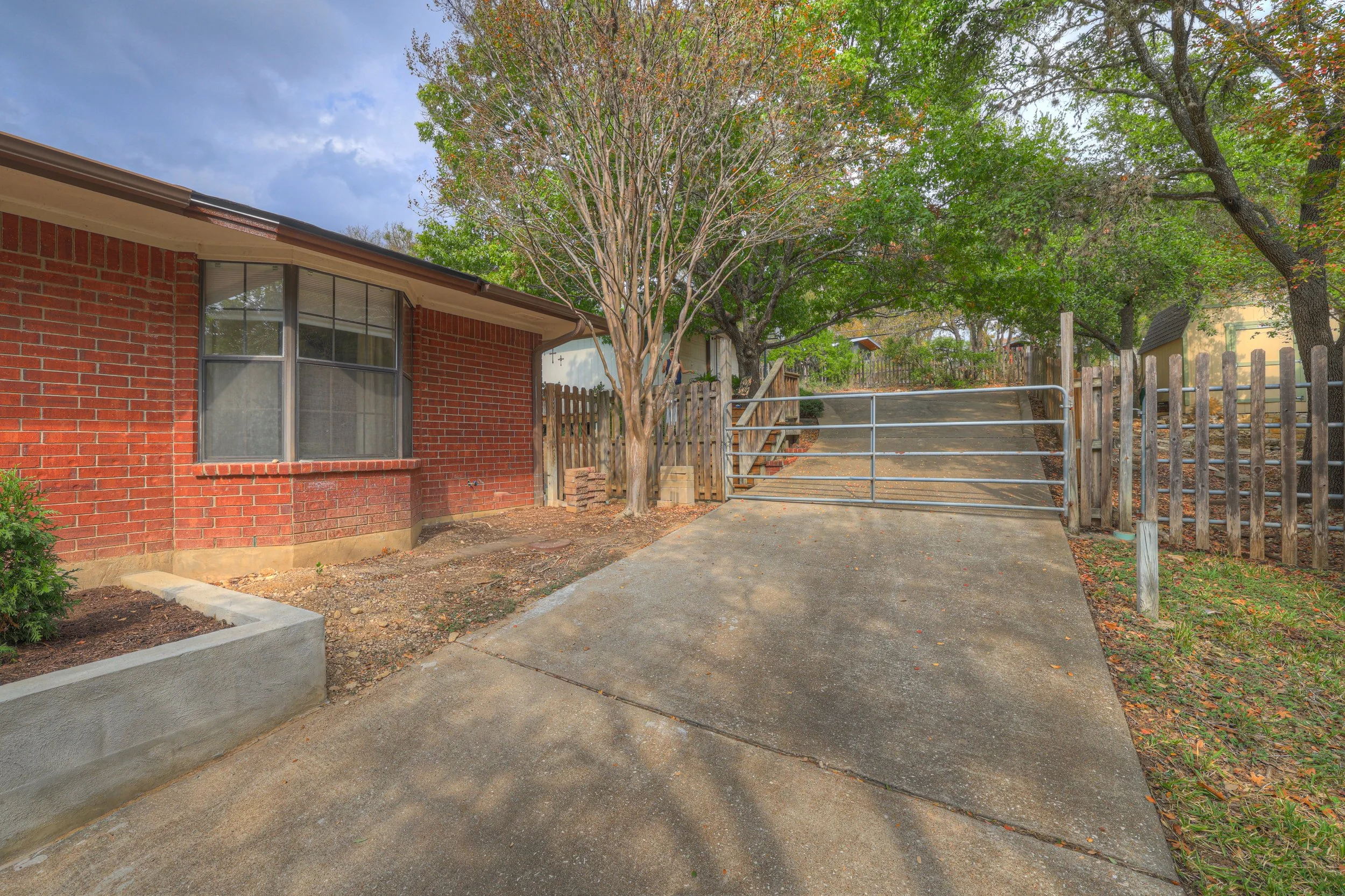 View of a brick house with a window, a tree, and a driveway with a metal gate and wooden fence, in a suburban neighborhood during daytime.
