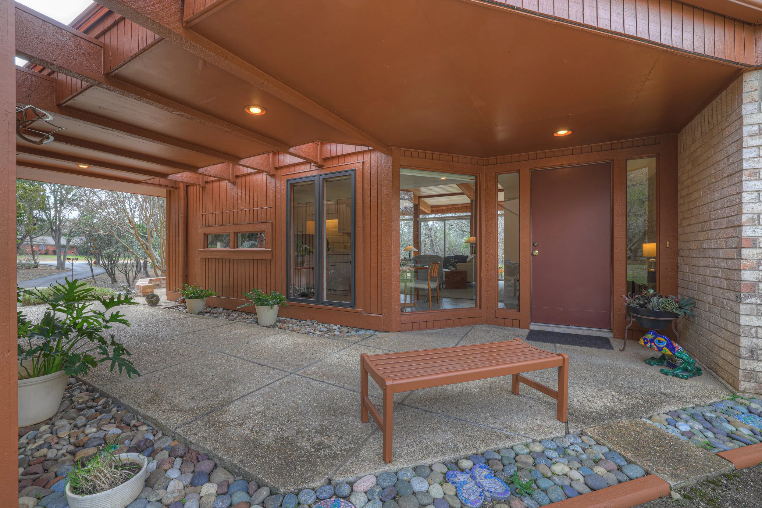 Covered patio area with potted plants, a wooden bench, and decorative stones, adjacent to a brick house with large glass windows and a door.