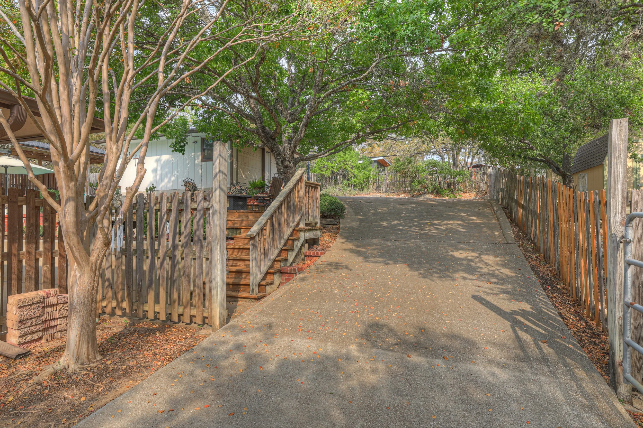 A driveway leading up to a house with a wooden staircase and railing, surrounded by trees and a wooden fence with fallen leaves scattered on the ground.