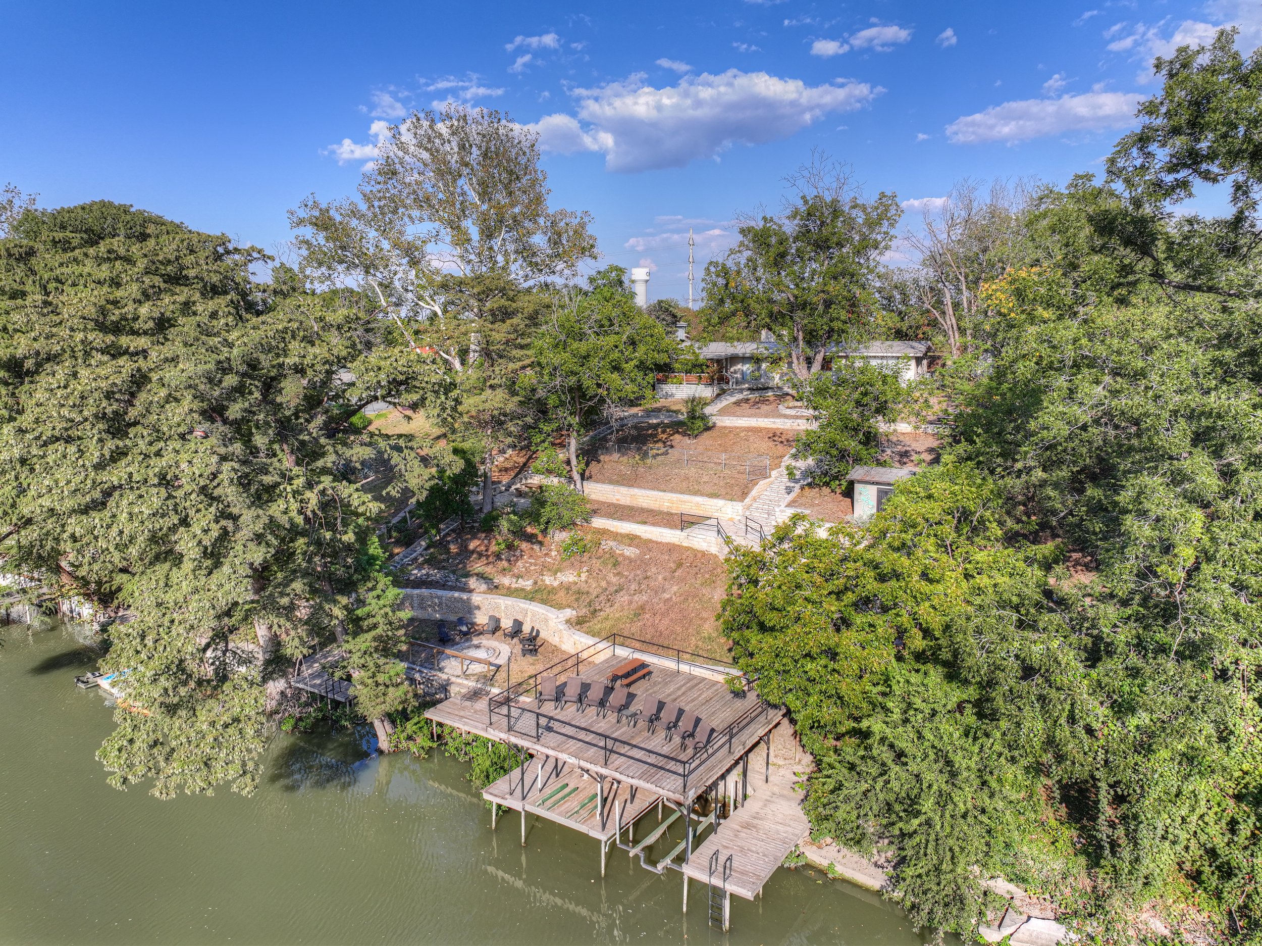 Aerial view of a lakeside backyard with a dock, outdoor seating, trees, and a house in the background.