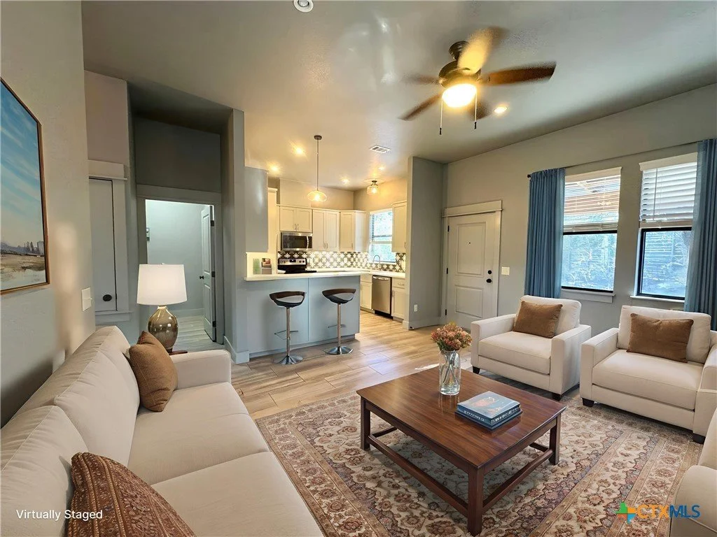 Living room with beige sofa and armchairs, wooden coffee table with a vase and books, area rug, windows with blue curtains, kitchen in the background with white cabinets and a breakfast bar with two stools, ceiling fan, and pendant lights.