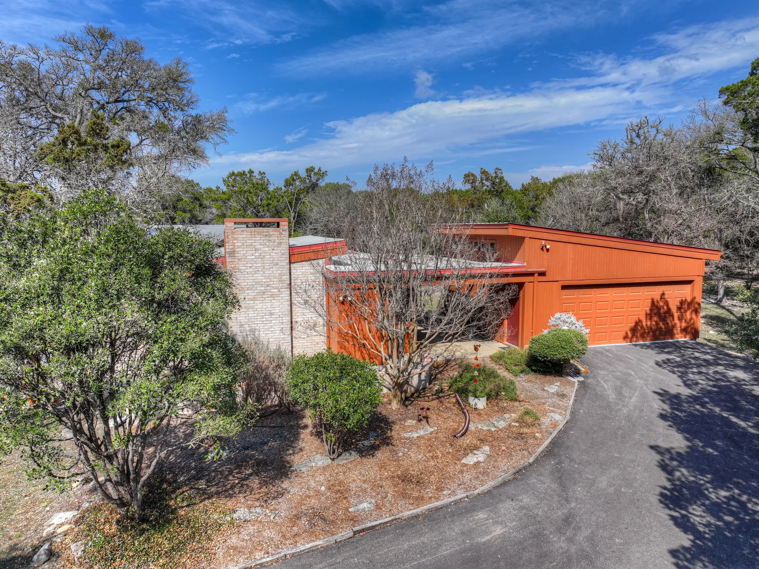 A mid-century modern house with an orange exterior, surrounded by trees and shrubs, under a partly cloudy blue sky.
