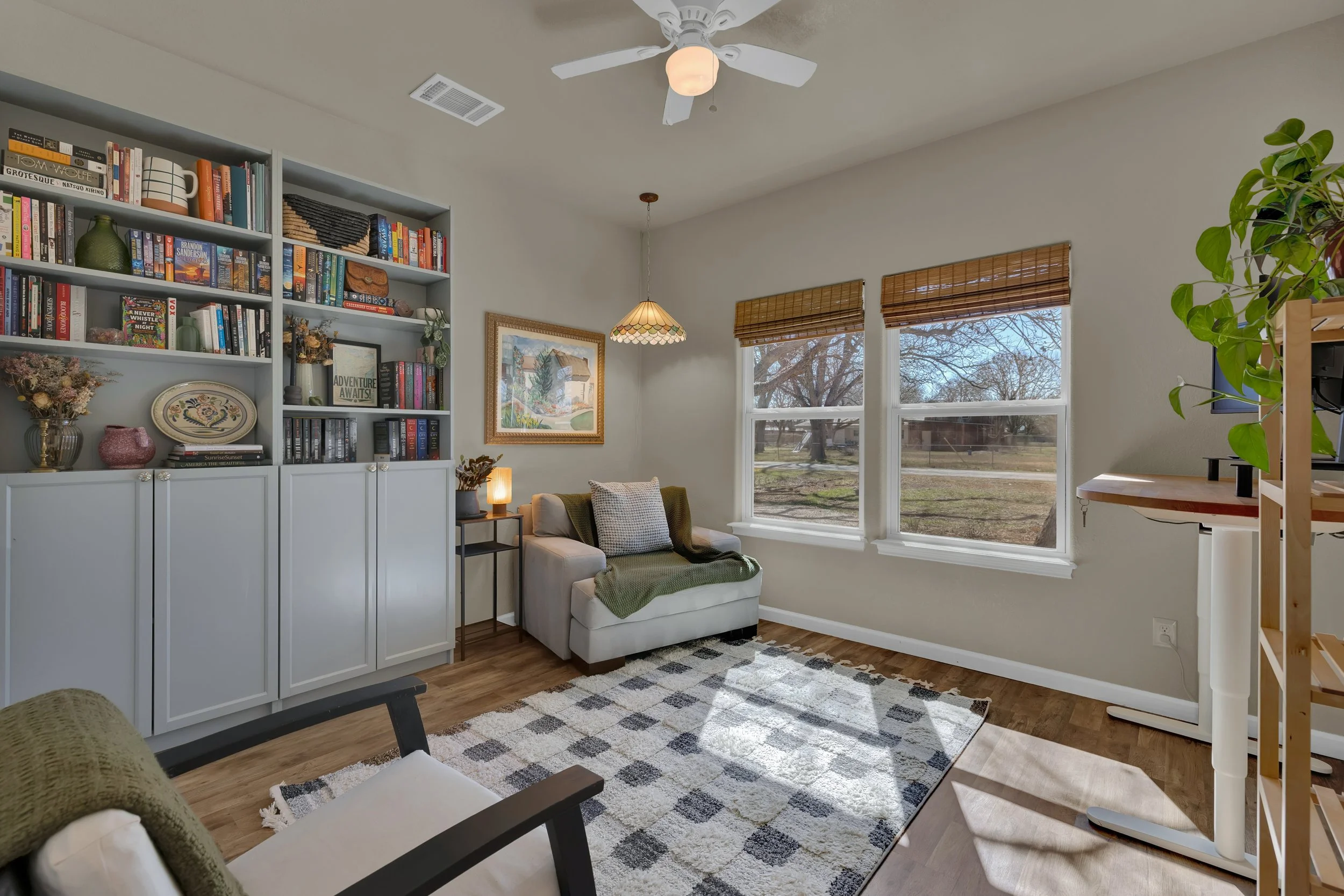 A cozy living room with large windows, a white bookshelf filled with books and decorative items, a small sofa with pillows, a patterned rug, and warm lighting from a table lamp and hanging light fixture.