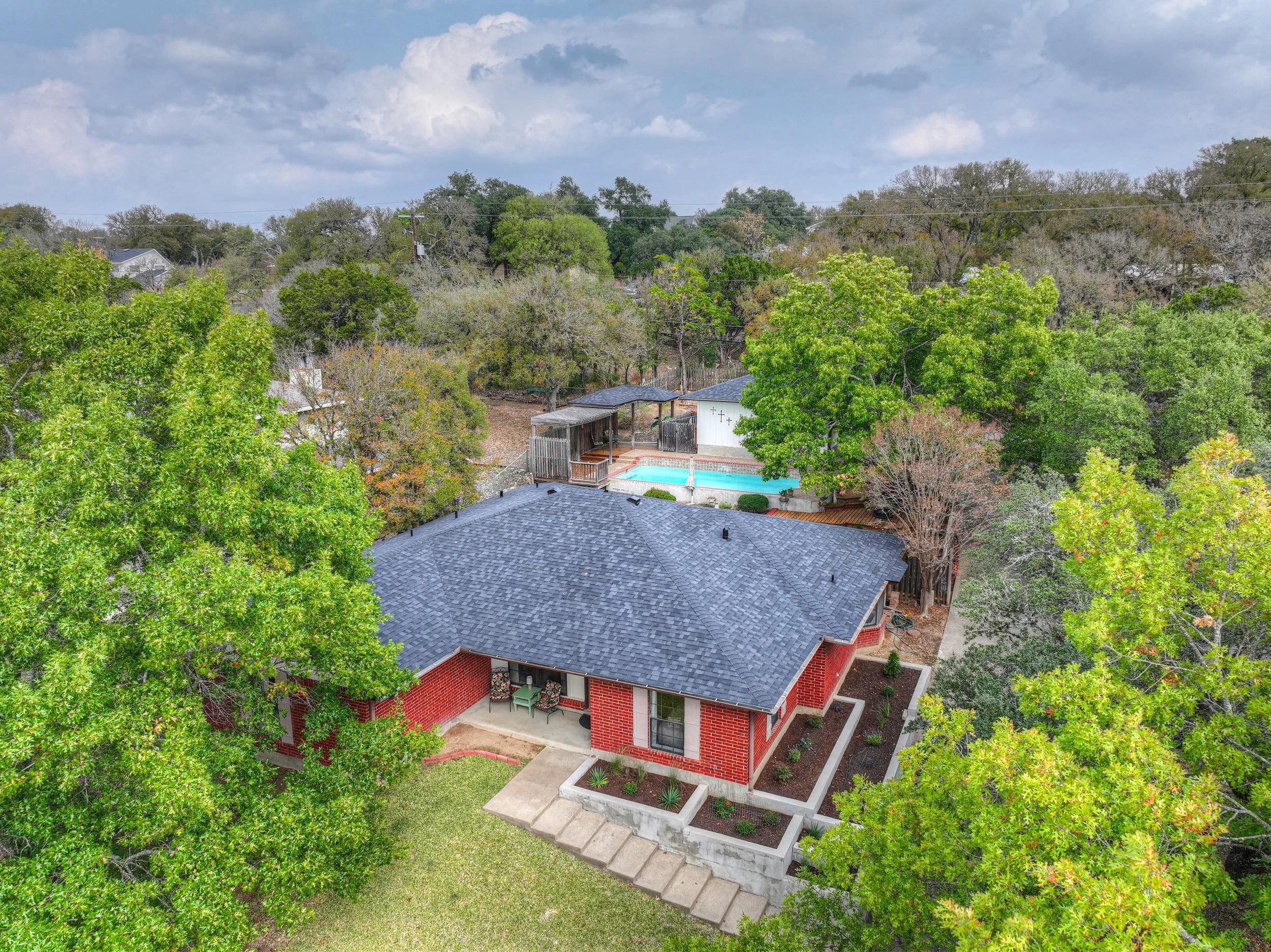 Aerial view of a house with a dark gray roof, brick walls, and a small front porch with outdoor furniture. The backyard features a swimming pool, pool deck, and a small wooden shed, surrounded by large green trees and a landscaped lawn.