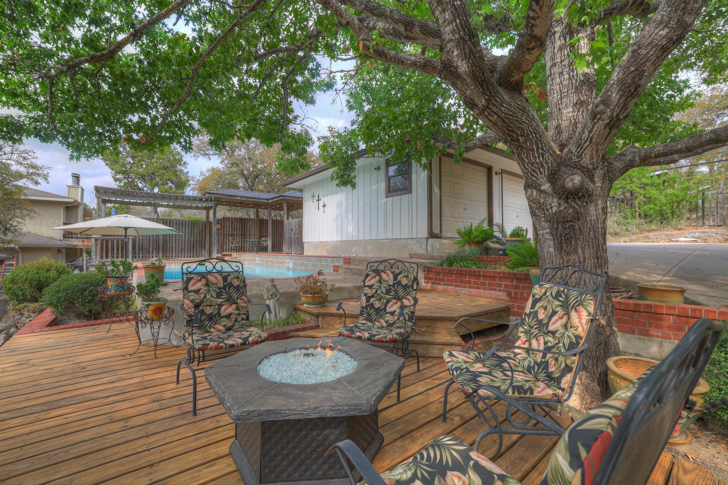 Backyard patio with wooden deck, a fire pit, four chairs with floral cushions, a large tree, a swimming pool with an umbrella, potted plants, and a small building with a garage door.