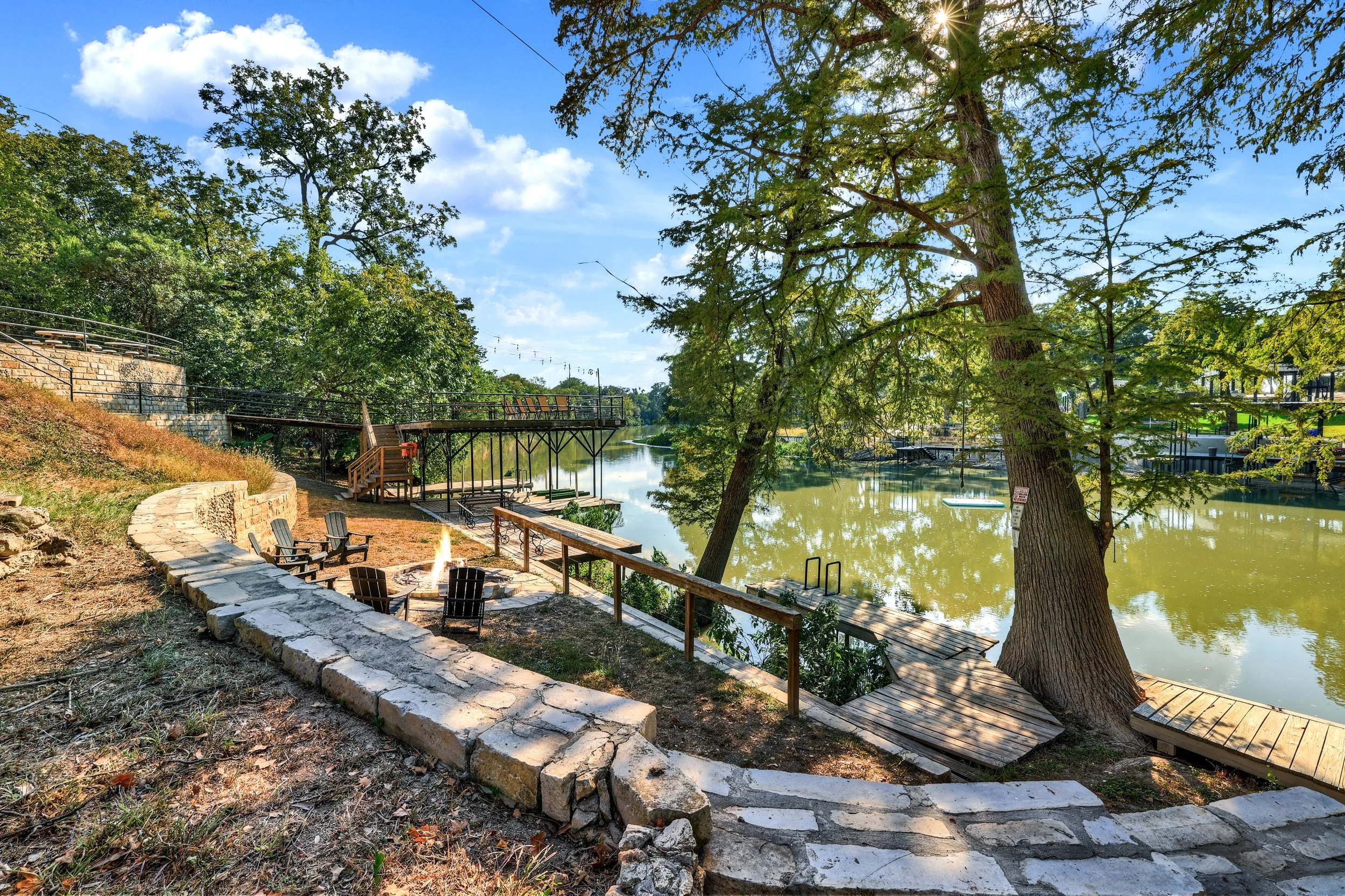 A lakeside outdoor scene featuring a firepit with chairs, trees, a wooden walkway along the water, and outdoor seating on a deck under a blue sky with clouds.