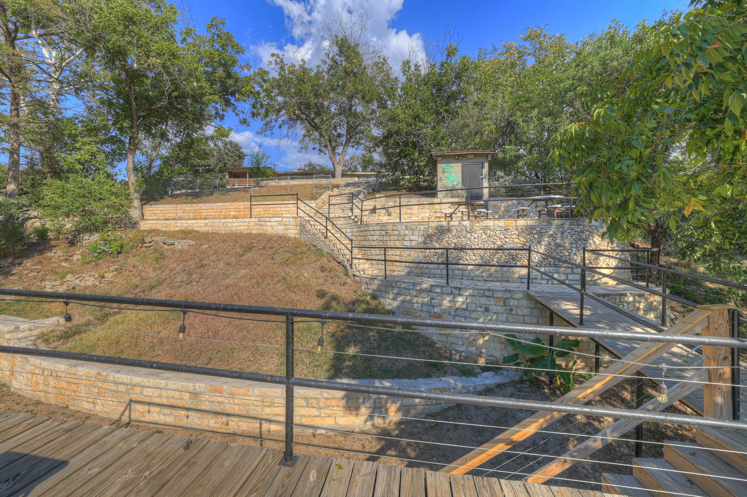 Multi-level stone and brick outdoor seating area with black metal railings, surrounded by trees and a clear blue sky.